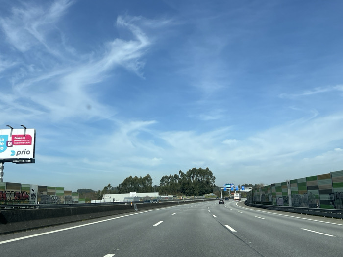 The image depicts a multi-lane highway under a clear blue sky with scattered cirrus clouds, located in Águas Santas, Portugal. The perspective is from within a vehicle traveling forward.

The paved highway features at least three visible lanes marked by white dashed lines, with a solid white line bordering the right shoulder. A dark gray concrete median barrier separates the opposing direction of traffic on the left.

On the left side of the highway, a large billboard displays an advertisement for "prio," featuring the Portuguese text "Preço ao posto, não no supermercado" on a blue, white, and red background. Beyond the billboard and the median, a long stretch of sound barriers, composed of rectangular panels in various shades of green, brown, and gray, is visible. These barriers exhibit extensive graffiti. Further behind the barriers, clusters of trees and a light-colored industrial-style building are present.

On the right side of the highway, another segment of similar paneled sound barriers runs parallel to the road, also showing signs of graffiti. Beyond these barriers, dense green foliage is visible.

Several vehicles, including passenger cars and at least one white commercial truck, are present on the highway ahead, traveling in the same direction as the camera. In the far distance, an overhead gantry with blue informational signs spans the highway. The scene is brightly illuminated by daylight. No individuals are visible outside of vehicles.