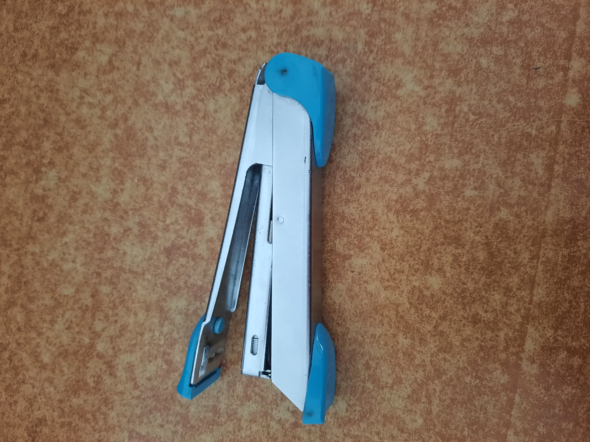 A blue and silver stapler is shown lying on a brown, textured surface. The stapler is open, with its upper arm lifted. The surface appears to be a table or countertop with a mottled, brown pattern. No people or other objects are visible in the image. The image does not provide any information about the location in Lhokseumawe, Indonesia.