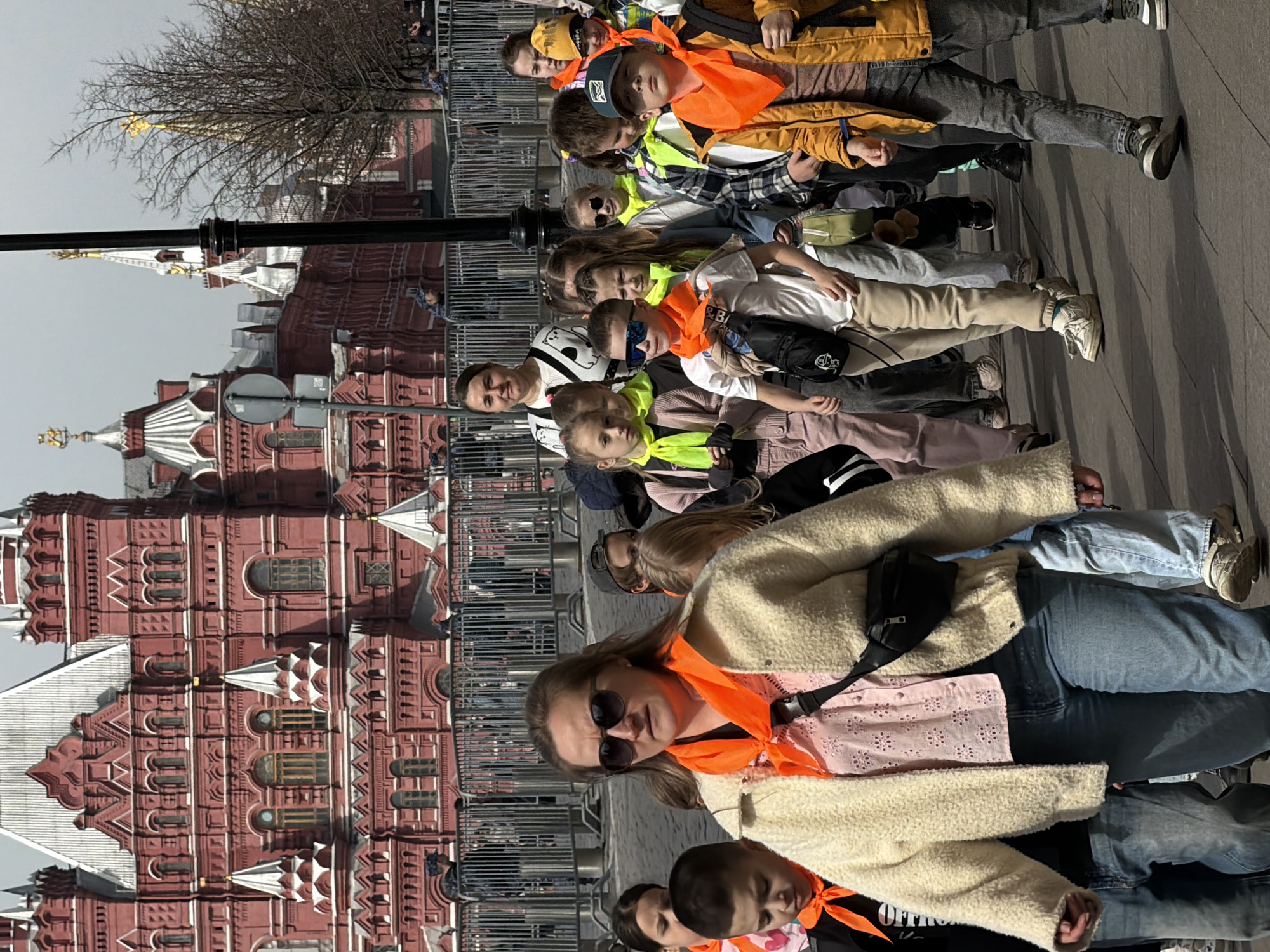 A group of people, primarily children, stand near a fence, with the State Historical Museum in the background. The museum is a large, red brick building. Most individuals wear brightly colored scarves, orange or yellow. The weather is overcast. A bare tree is visible in the background, as well as light posts. The location is Moscow, Russia.