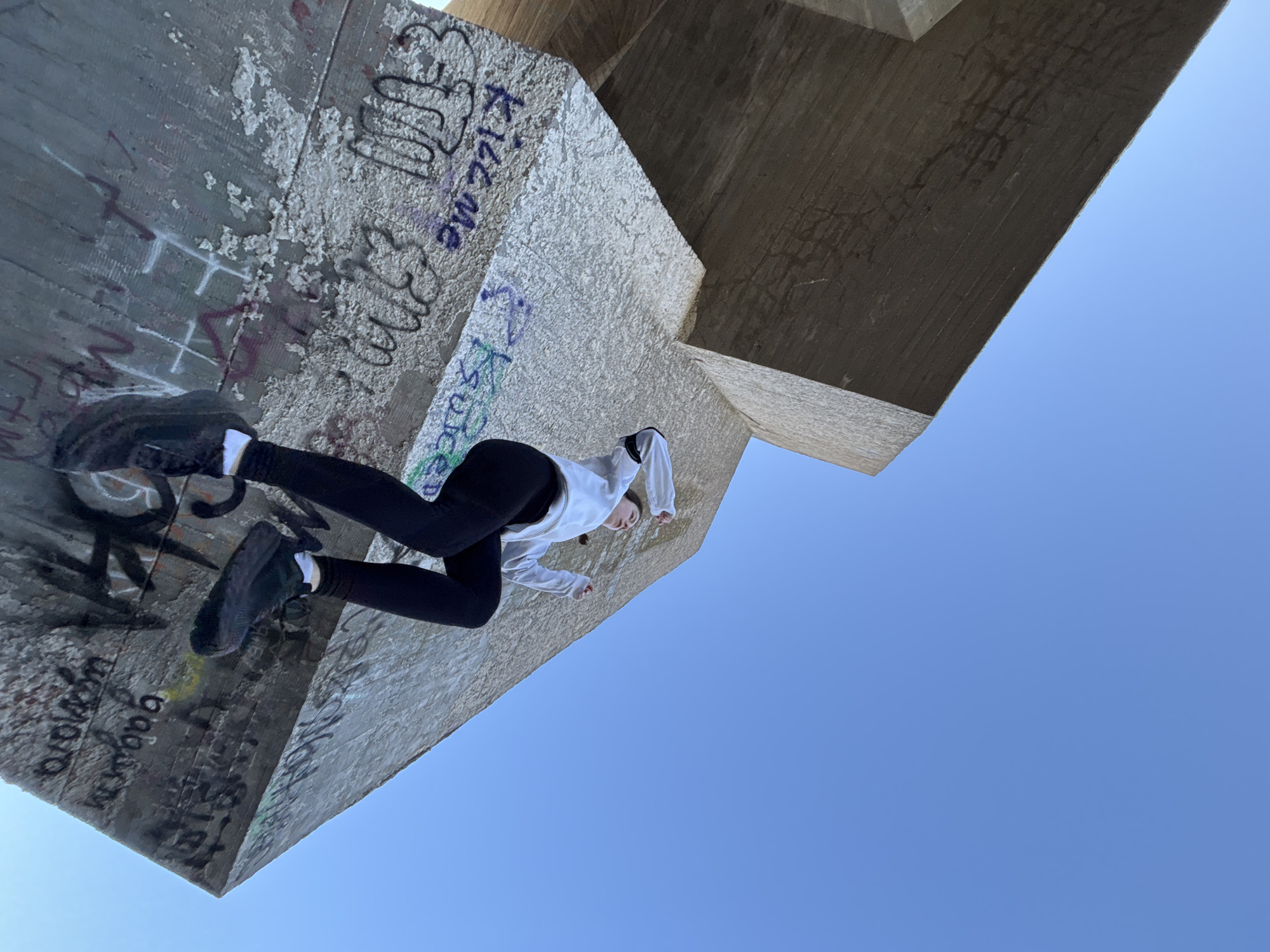 This image, taken from a low angle, features a young person performing an optical illusion or an act of parkour on a large concrete structure under a clear blue sky. The subject, likely female, is dressed in black leggings, a white hooded long-sleeved top, and dark sneakers with white socks. She is positioned horizontally across the frame, with her legs bent as if running or posing on a vertical wall, creating the impression that she is defying gravity.
The setting consists of a massive, textured concrete structure, possibly an underpass, bridge support, or a brutalist architectural element, typical of urban environments like Varna, Bulgaria. This concrete surface is extensively covered in graffiti and paint. Visible text includes "Kiss me" in English, and several Cyrillic writings such as "pizda" and "Gabava," alongside numerous other illegible tags and scribbles in various colors.
The background is a vast expanse of bright, clear blue sky, indicating excellent weather conditions and a daytime setting, likely midday or early afternoon due to the even, bright lighting. There are no clouds or obstructions in the sky. The overall scene conveys a sense of youthful daring or artistic play within a rugged, urban landscape marked by human interaction and decay.