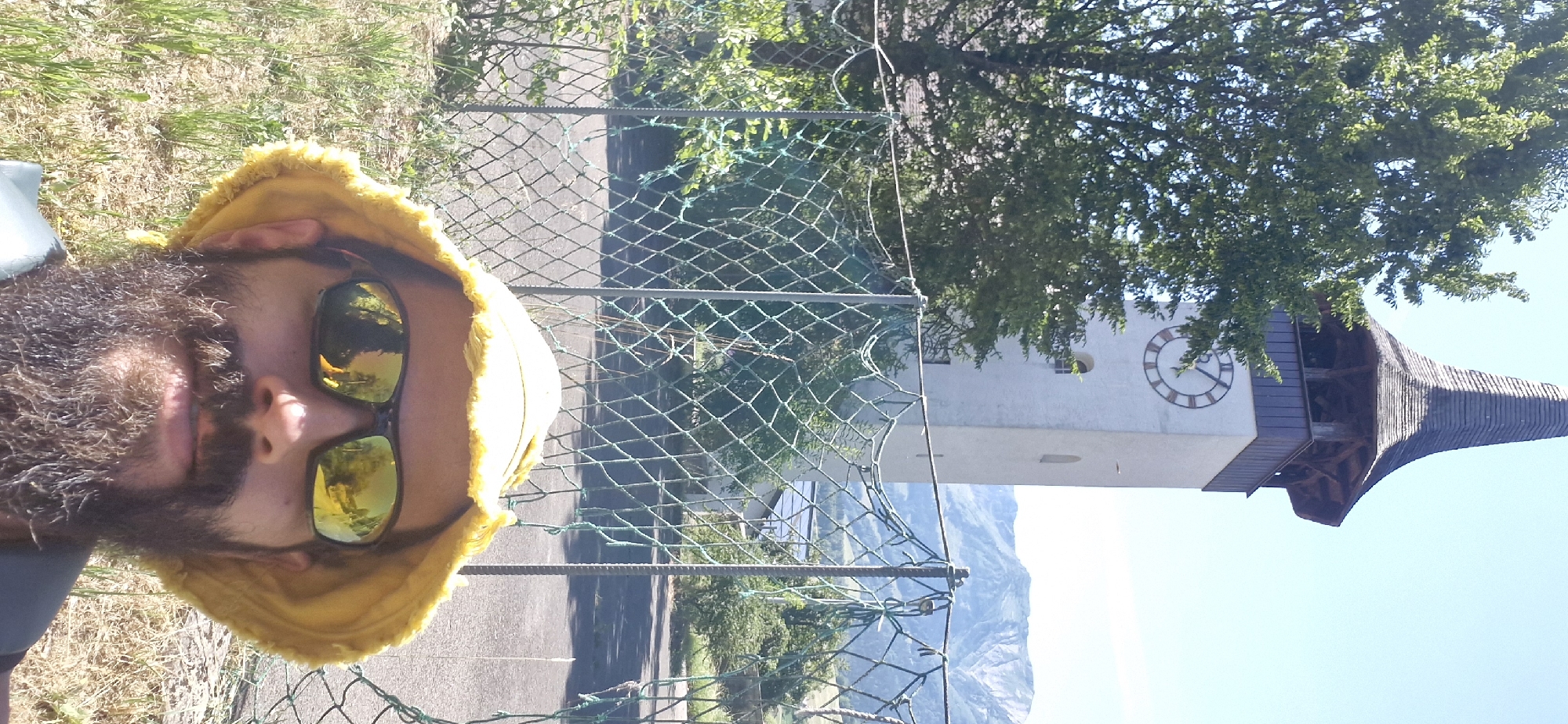 This is a selfie taken by a man with a beard and sunglasses. He is wearing a yellow bucket hat. The background features a white clock tower with Roman numerals on its face, a dark, pointed roof, and lush green trees. Mountains are visible in the distance under a clear blue sky. A chain-link fence runs across the foreground. The image appears to have been taken outdoors on a sunny day, possibly in the morning or afternoon. The man's expression is relaxed and happy. There is no visible text in the image. The scene is likely Wattenwil, Switzerland.