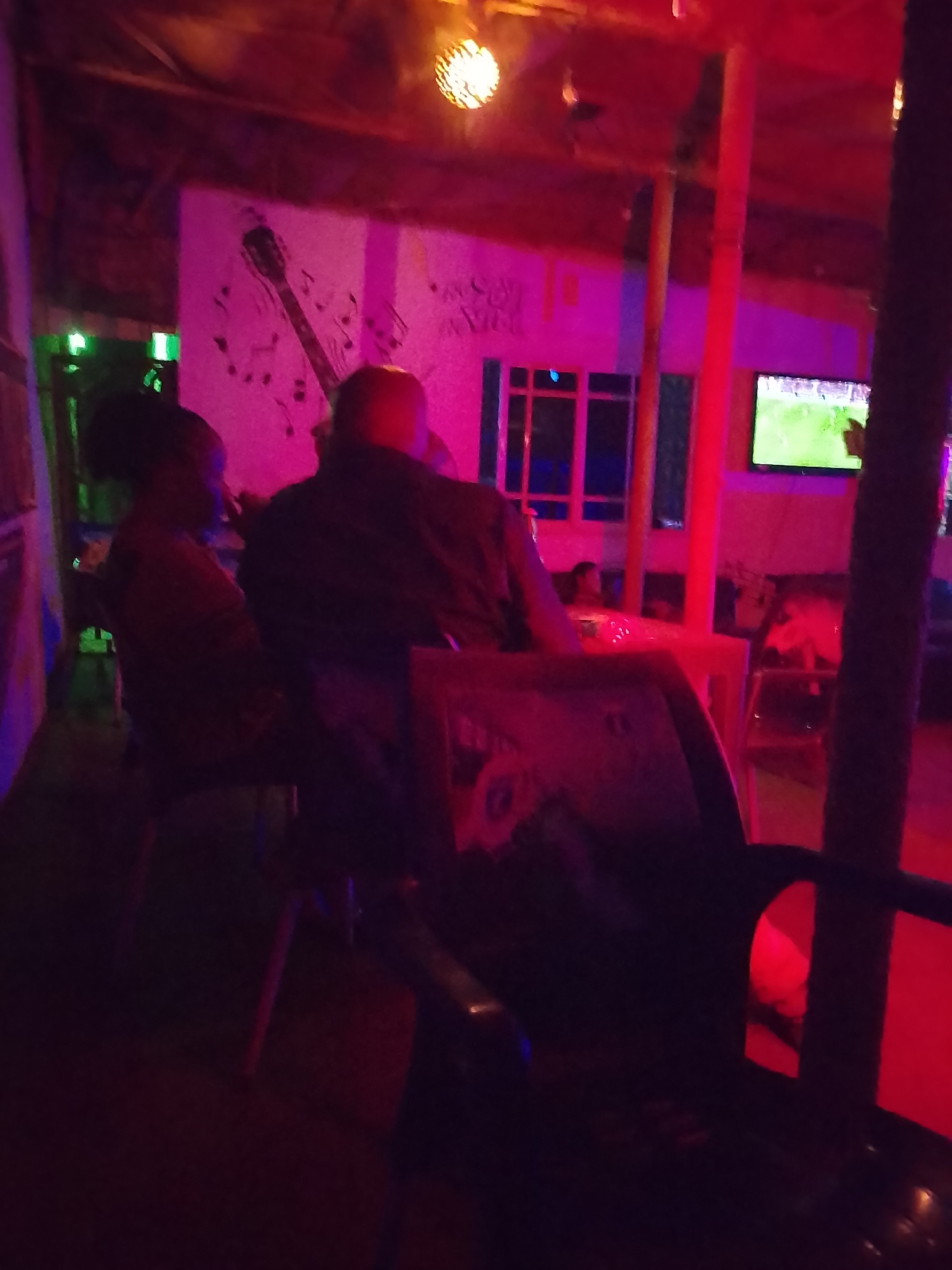Location: Likely a bar or restaurant in Yelwa, Nigeria.
Scene: Two individuals are seated at a table indoors. A person with dark hair is facing left, while another, with a lighter-colored head, is facing away from the camera. The scene is illuminated by colored lighting, primarily red and purple.
Objects: A guitar and musical notes are painted on the wall. A window with a grid pattern and a television displaying a green screen (possibly a sports game) are visible in the background. A decorative light fixture hangs from the ceiling. Chairs and a table are present.
Environment: The space appears to be enclosed. The lighting is dim and colorful, suggesting an evening setting.
