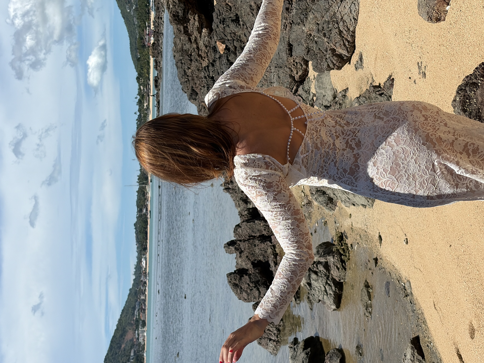 A woman stands facing away from the camera on a sandy beach in Baan Bo Phut, Thailand, with her arms outstretched. She is wearing a white, long-sleeved lace dress with a low back adorned with pearl beading. The beach is rocky, with dark volcanic rocks scattered along the shore. The sea is calm, with a few sailboats visible in the distance. Lush green hills rise behind the coastline, dotted with buildings. The sky is partly cloudy, with dramatic formations of white clouds against a blue expanse. The sun appears to be shining, casting shadows on the sand and rocks.