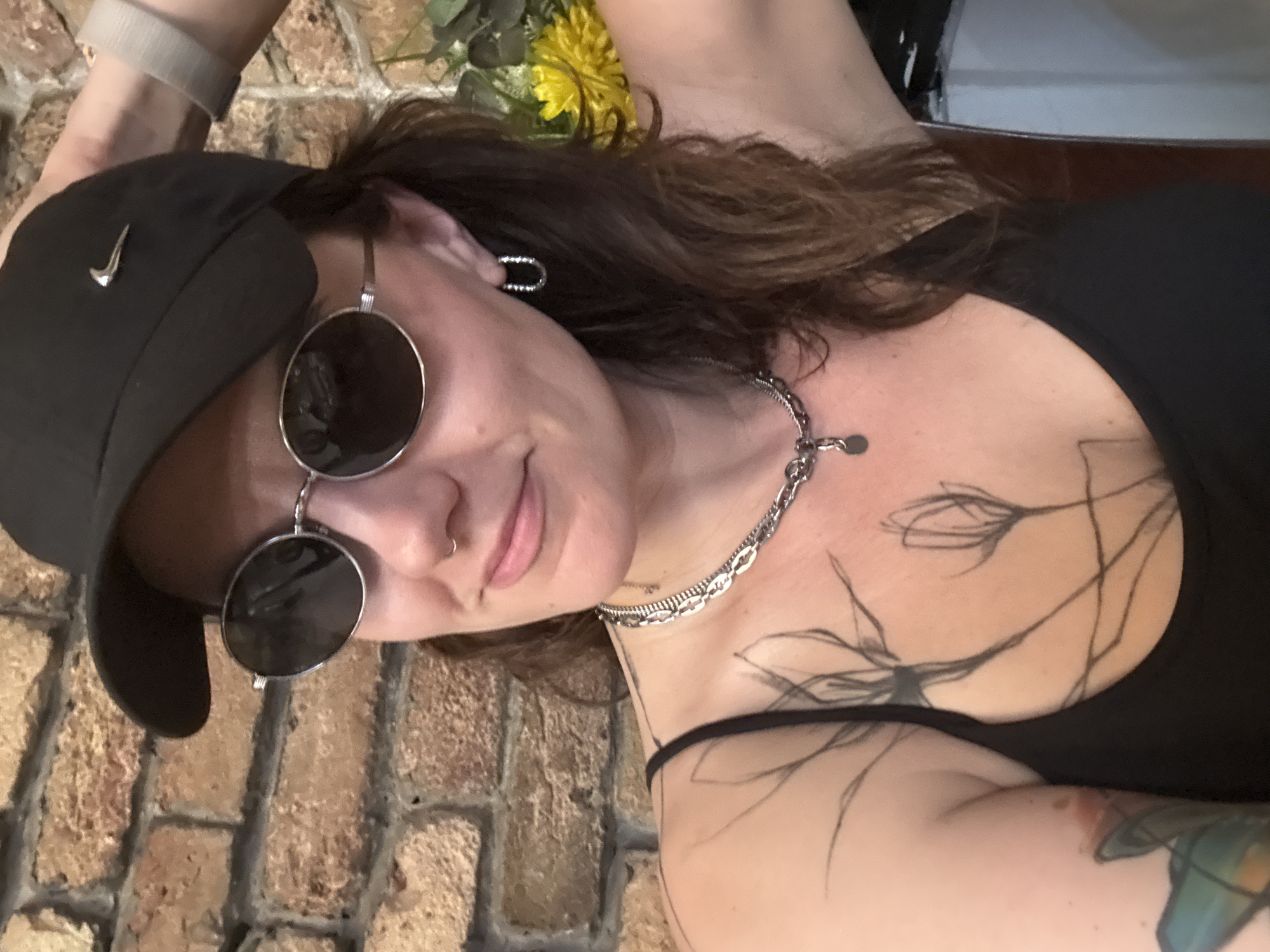 A woman wearing a black Nike baseball cap, oval sunglasses, and a black tank top is shown in a selfie. She has dark brown hair and is wearing a silver chain necklace and hoop earrings. Tattoos are visible on her chest and arm. The background is a brick wall, and a yellow flower arrangement is visible to the right. The scene is indoors, and the lighting is moderate. The location is not specified in the image itself.