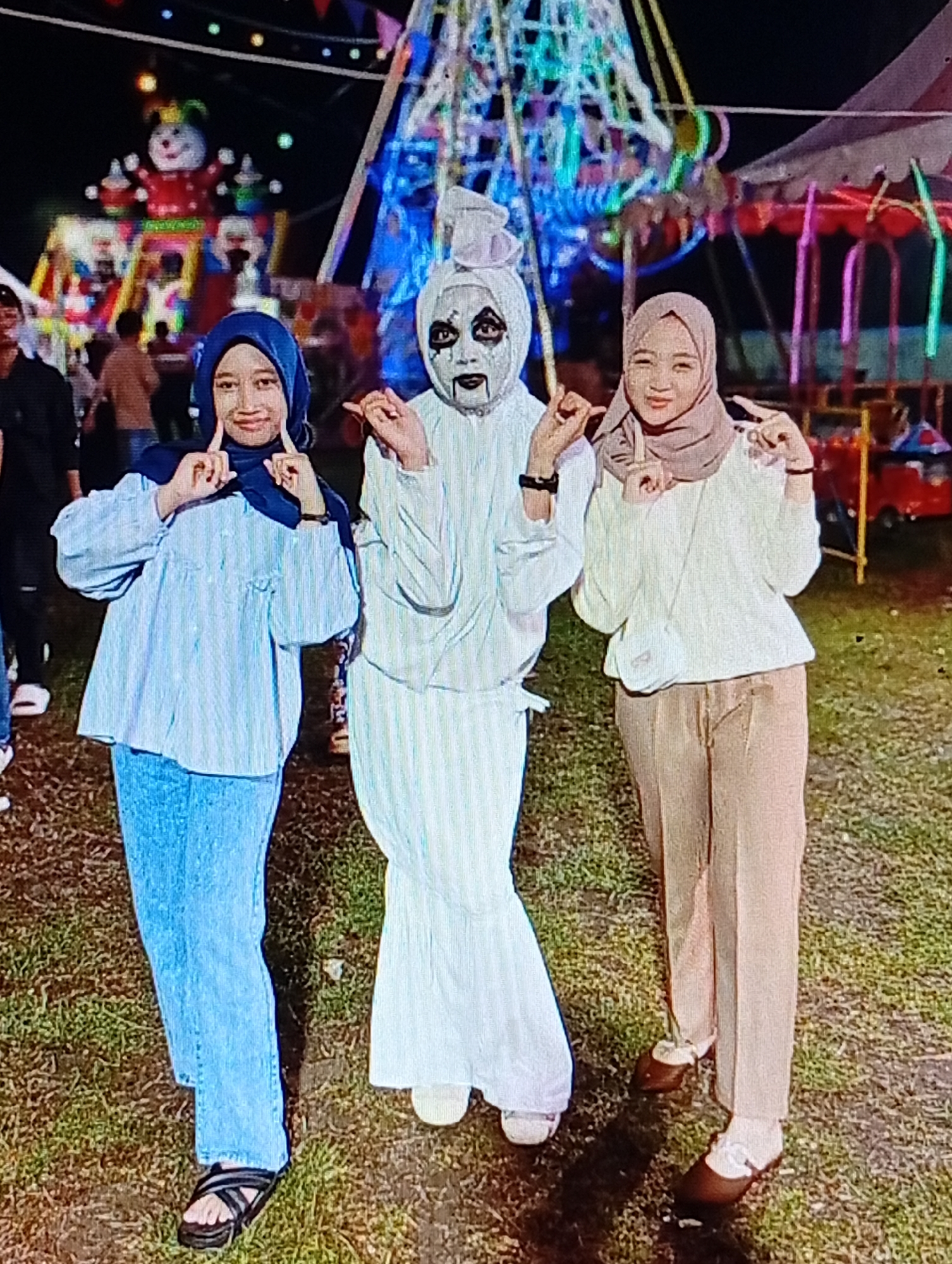 The image depicts three females posing outdoors at night against the illuminated backdrop of a fairground or carnival located in Lhokseumawe, Indonesia.
From left to right:
1. A female with light skin is wearing a dark blue hijab, a light blue long-sleeved top with a subtle textured or striped pattern, light blue jeans, and black strap sandals. She is smiling at the viewer and making a "finger heart" gesture with both hands positioned near her cheeks.
2. A central female figure is dressed in a full white costume, including a white head covering resembling a turban or draped cloth, a loose-fitting white long-sleeved top, and wide-leg white pants. Her face is covered in white makeup with distinct black markings around the eyes, an elongated black nose outline, and black lines extending from the corners of her mouth. She is wearing white flat shoes. She is also making a "finger heart" gesture with both hands positioned near her face, displaying a subtle smile or smirk.
3. A female with light skin is wearing a light brown or beige hijab, a light cream-colored long-sleeved top, light brown wide-leg trousers, and dark brown flat shoes with visible white socks. A small, light-colored crossbody bag is slung across her body. She is smiling at the viewer and making a "peace sign" or "V-sign" gesture with both hands positioned near her face.
The individuals are standing on a ground surface composed of grass with visible patches of bare earth. The background features various brightly lit attractions typical of a night fair. To the mid-left, a large, colorful, illuminated structure resembling a snowman or clown with a jester hat (primarily red, white, and green) is visible, surrounded by other smaller, colorful lights. Further to the right in the background, a tall structure, possibly a Ferris wheel, is illuminated with blue and white lights. To the far right, part of a carousel or similar ride with red, pink, and green elements, a small red car ride, and a white tent structure are visible. The scene is brightly lit by the artificial lights of the fair attractions against the dark night sky.