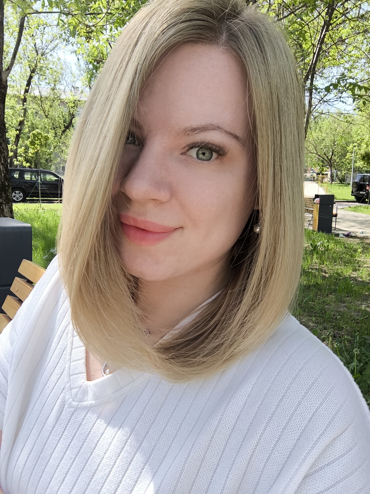 A woman with blonde hair and green eyes is sitting on a bench outside. She is wearing a white shirt and a silver necklace. In the background, there is grass, trees, a black car, and a trash can. This location is in Fryazino, Russia.