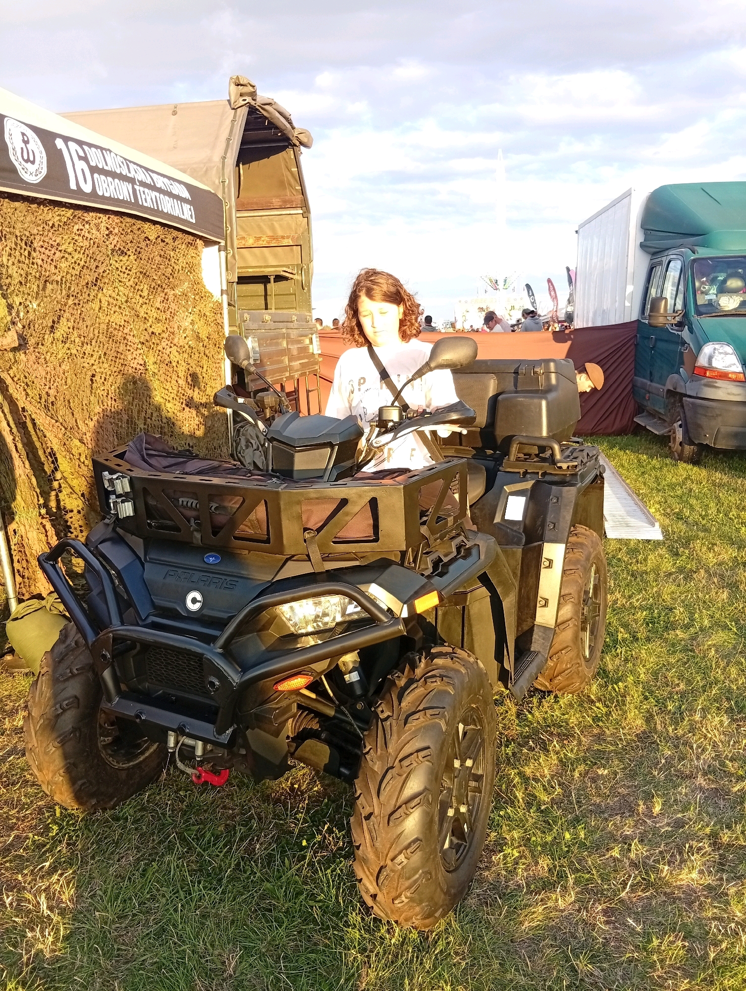 The image captures an outdoor scene on a grassy field, likely at an event in Szewce, Poland, given the text on the banner. In the foreground, a dark green Polaris all-terrain vehicle (ATV) is parked. A young person with wavy, light brown hair is seated on the ATV, wearing a light-colored t-shirt and a dark strap across their chest. They are looking down, with their face partially obscured.
To the left of the ATV, a large tent with a banner that reads "16 DOLNOŚLĄSKA BRYGADA OBRONY TERYTORIALNEJ" is visible. The tent is draped with camouflage netting. Behind the ATV and the tent, a military-style truck with a canvas cover is parked. Further in the background, other tents, people, and a green van are present. The sky is bright with scattered clouds, and the sunlight casts long shadows on the grass, indicating it is late afternoon or early evening. The overall setting appears to be an open-air event or exhibition.