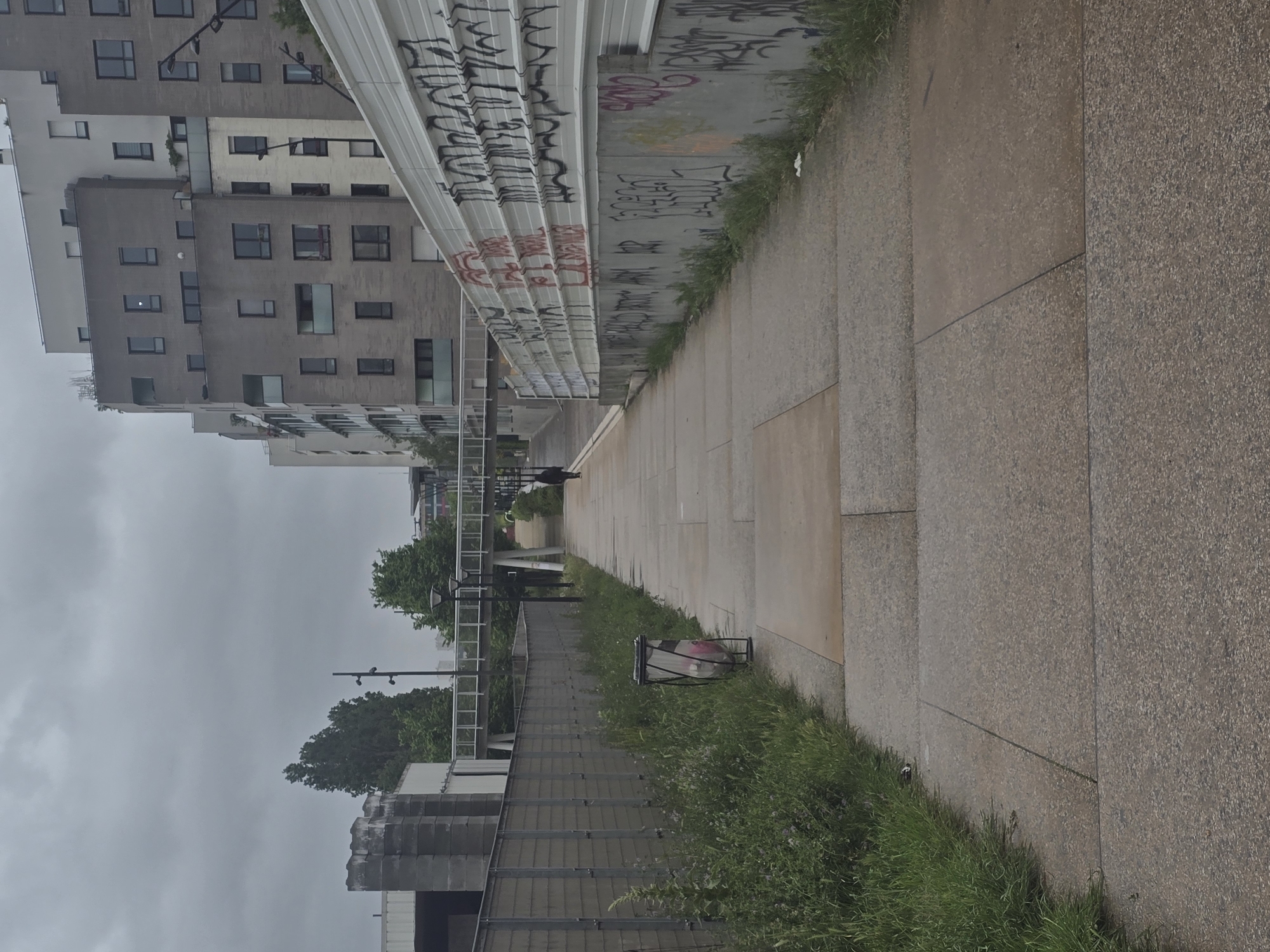 The image captures a street scene in Aubervilliers, France, characterized by modern architecture and urban elements. A sidewalk, composed of large square pavers, dominates the right side of the frame, bordered by a strip of grass. A trash can sits on the grass next to the sidewalk. A fence runs parallel to the sidewalk, separating it from a lower area with trees and a pedestrian overpass. On the left, tall apartment buildings with multiple windows rise into a gray, overcast sky. A corrugated metal structure overhead is covered in graffiti. In the distance, a figure is walking on the sidewalk.