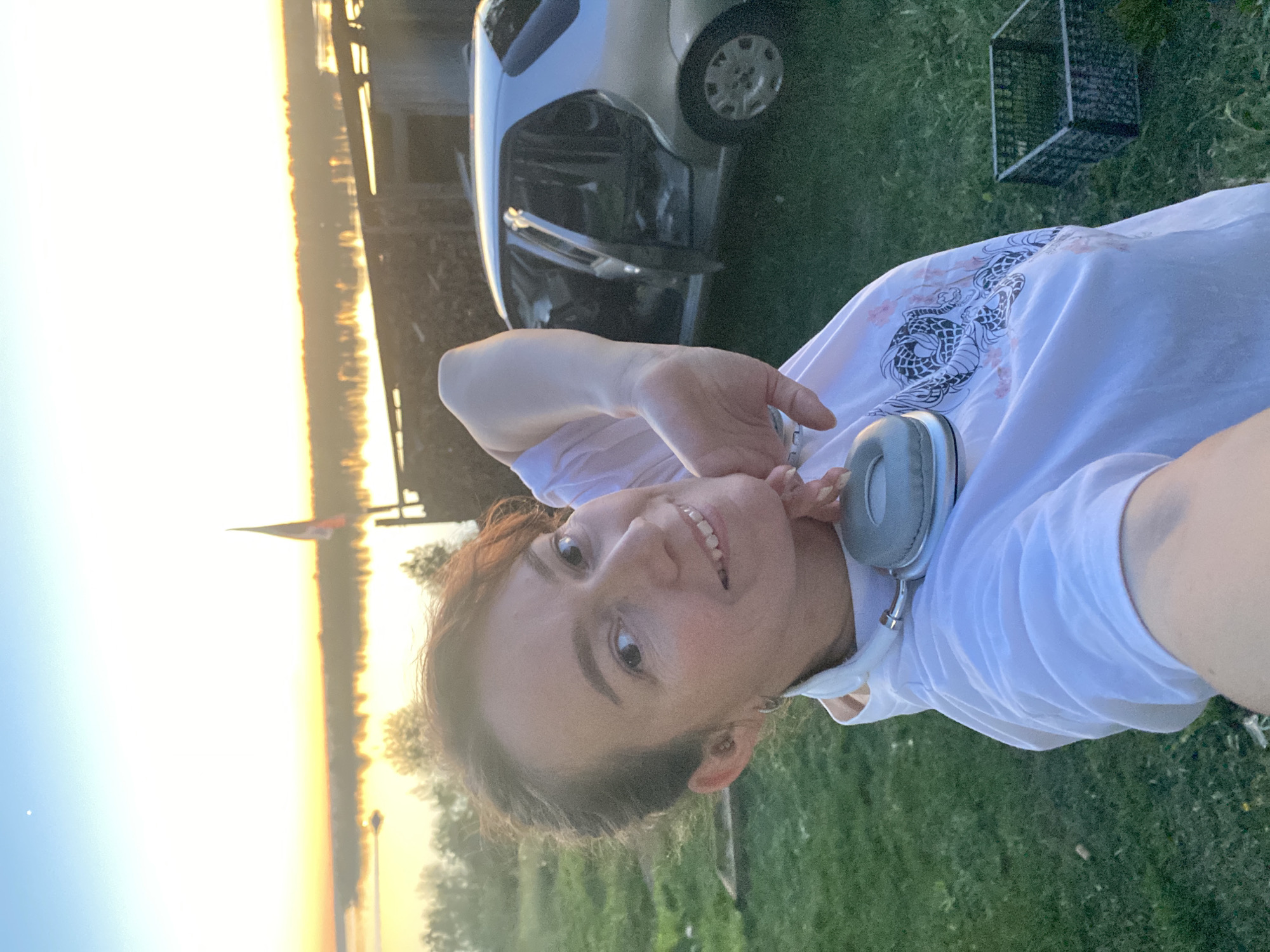 A woman is lying on grass in an outdoor setting with a body of water and a car visible in the background. The water reflects the sunset. The woman is wearing a white t-shirt with a dragon design and headphones around her neck. A wire mesh container is in the background. The location is Ольховка, Russia.