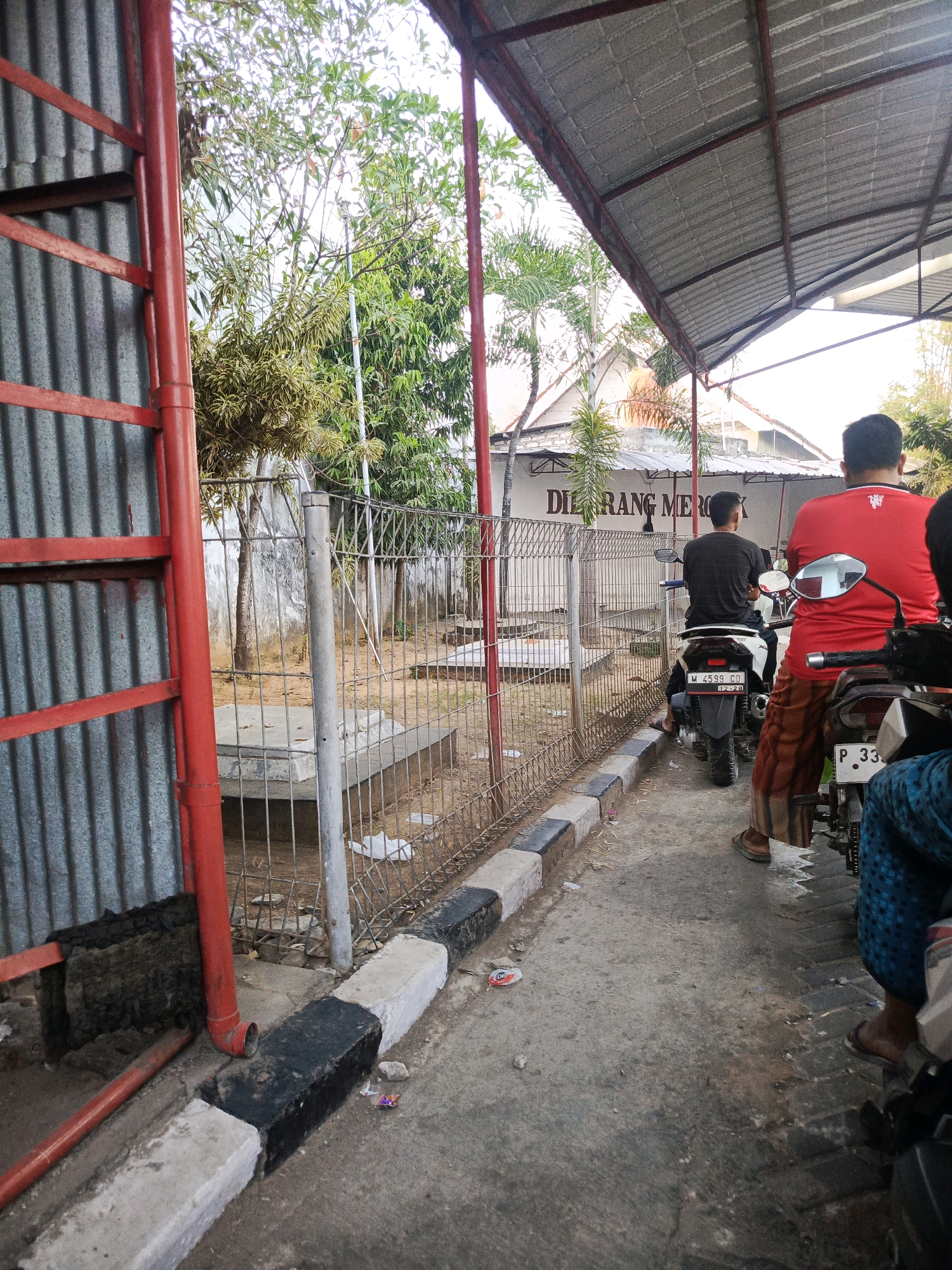The image shows an outdoor scene, possibly a parking area or walkway, with a corrugated metal fence on the left and a metal roof structure extending overhead. In the middle ground, a wire fence encloses an area with some vegetation and what appear to be concrete structures. A sign in the background reads "DILARANG MEROKOK," which translates to "NO SMOKING" in Indonesian. There are at least three people visible, all on or near motorcycles. One person in a black shirt is riding a white motorcycle. Another person in a red shirt is sitting on a motorcycle, facing away from the camera. A third person, partially visible in the foreground, is also on a motorcycle. The ground is paved with concrete. The environment appears to be in a warm climate, with trees and bright daylight. The location is identified as Gladak Anyar, Indonesia.