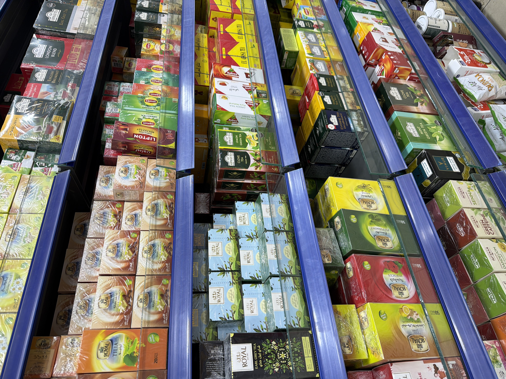 The image shows a close-up, eye-level view of shelves stocked with various brands and types of tea boxes in a retail setting, likely a supermarket or specialty store. The shelves are a vibrant blue color, and the products are predominantly packaged in colorful cardboard boxes, many with cellophane wrapping. Brands visible include Royal, Lipton, Ahmad Tea, and others that are less clear due to resolution or packaging design. The teas are of various flavors and types such as Earl Grey, Ginger & Cinnamon, Green Tea, Lemon, Hibiscus, and Peppermint. Some boxes are stacked vertically, while others are arranged horizontally. The lighting appears to be bright, illuminating the products clearly. The environment is clean and organized, typical of a well-maintained retail display. No people are visible in the frame. The scene is static, focusing solely on the product display. The image appears to be taken in Sharm El Sheikh, Egypt, judging by the presence of Arabic text on some of the packaging.