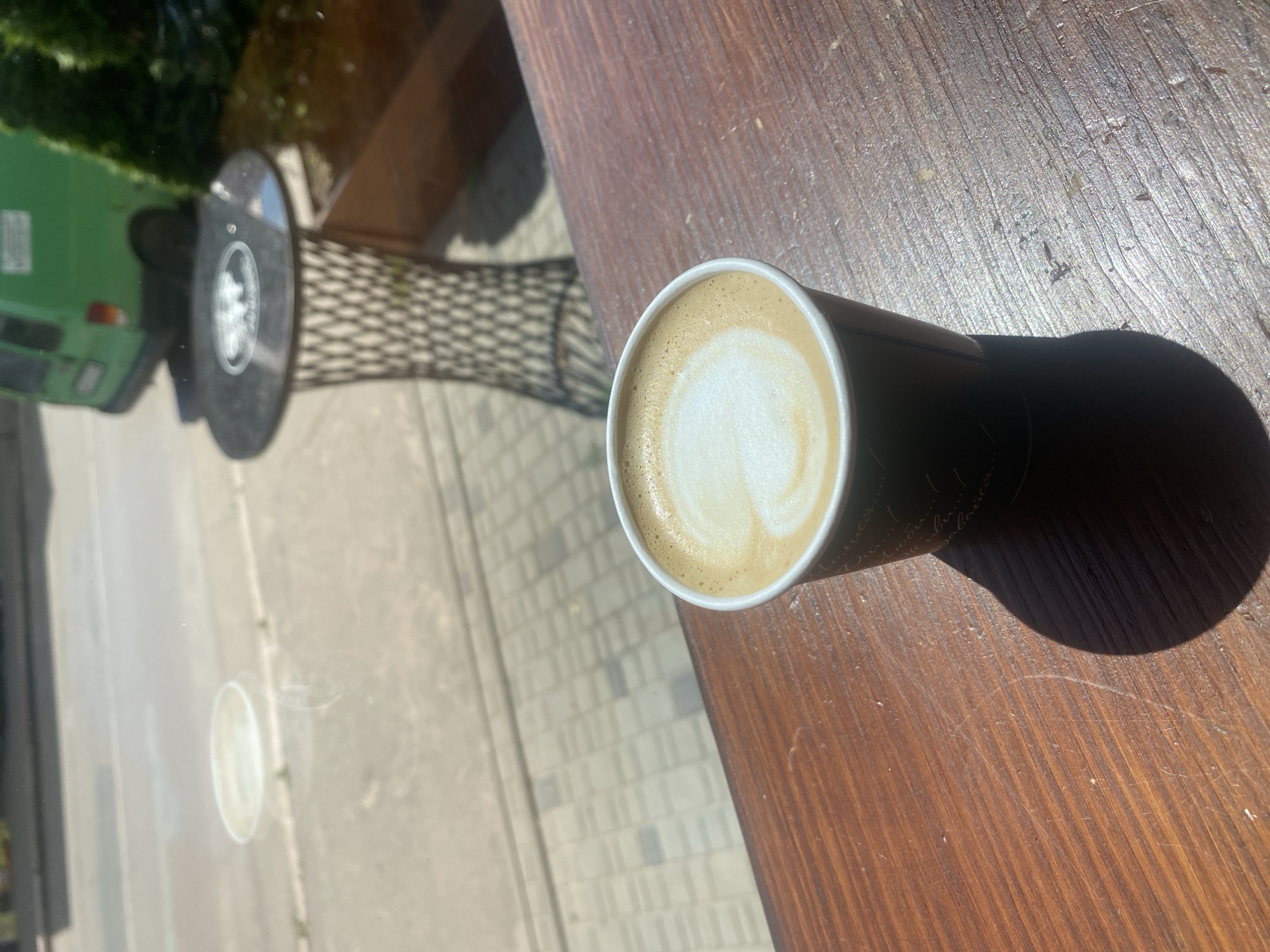 A latte in a dark paper cup sits on a wooden table, bathed in sunlight. The latte has a heart shape in its foam, a classic sign of latte art. The table has a natural wood grain texture, with some scratches and debris visible, indicating it might be an outdoor table.
In the background, seen through a glass pane, is an outdoor scene. A green vehicle, possibly a van or small truck, is partially visible, parked on a paved area. The ground appears to be a sidewalk or patio made of light-colored bricks or pavers. There's also a glimpse of a metal table with a patterned surface, suggesting an outdoor seating area. Trees with lush green foliage are visible further in the background. The overall impression is that of a cafe or outdoor seating area on a sunny day. There is no visible text. The scene is likely captured in Khoroshiv, Ukraine. The bright sunlight and clear background suggest it is daytime.