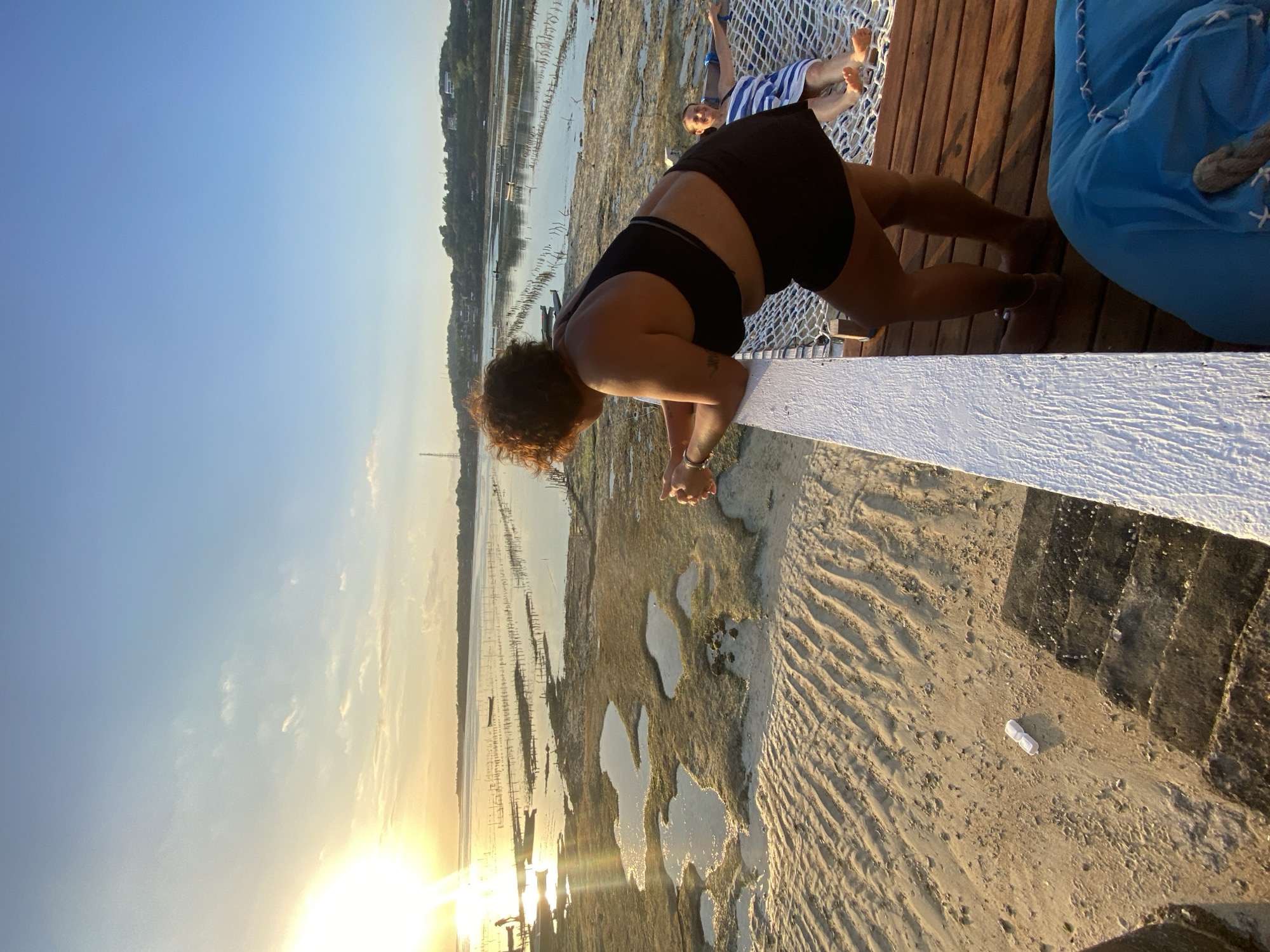 A medium shot captures two people on a hammock over a sandy beach with shallow water and wooden posts. The person in the foreground, a woman with curly hair wearing black shorts and a black top, leans over the edge of a wooden structure, her arms extended. Her gaze is directed downwards. In the background, another person, partially visible and wearing striped shorts, reclines on the hammock.
The scene appears to be set during a sunset, indicated by the warm glow and the low angle of the sun visible in the lower left corner of the frame. The sky is clear blue with hints of light clouds. The ground is a mix of sand and shallow water, with many wooden posts jutting out from the water, suggesting an oyster or seaweed farm. The overall atmosphere is relaxed and serene. The image is taken from a slightly elevated perspective, looking down towards the beach and water. No visible text is present in the image.