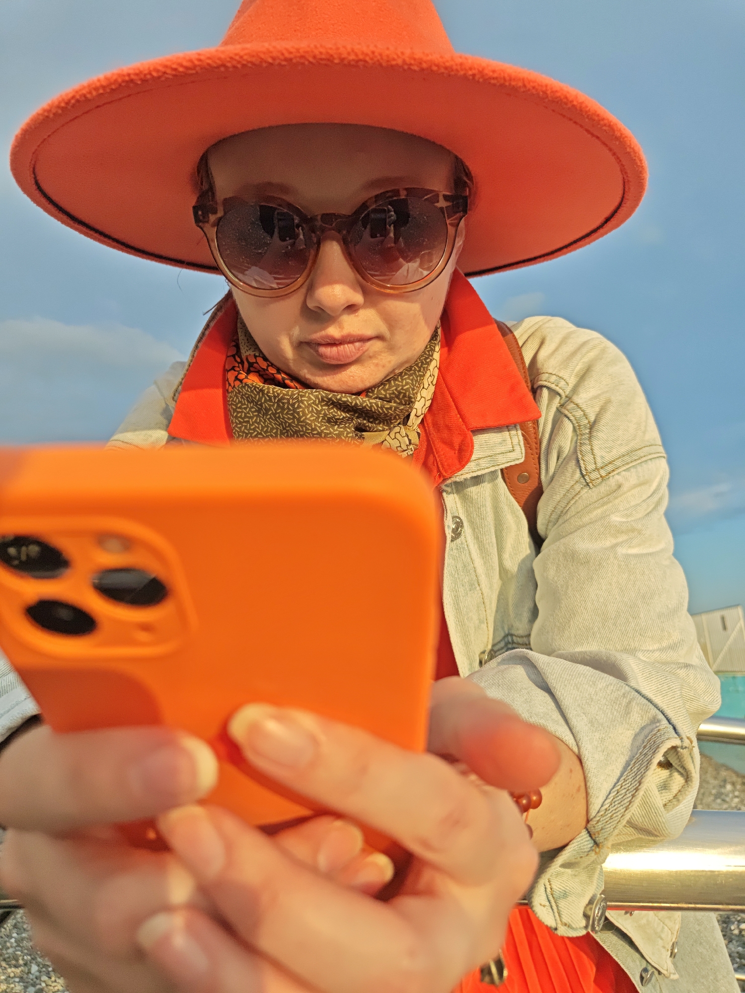 A person wearing a bright orange hat and sunglasses is looking at a smartphone in an orange case. The person is also wearing a denim jacket over an orange top, with a patterned scarf around their neck. The background shows a blue sky and a railing, suggesting an outdoor location. The photo was taken in the city of Sochi, Russia.