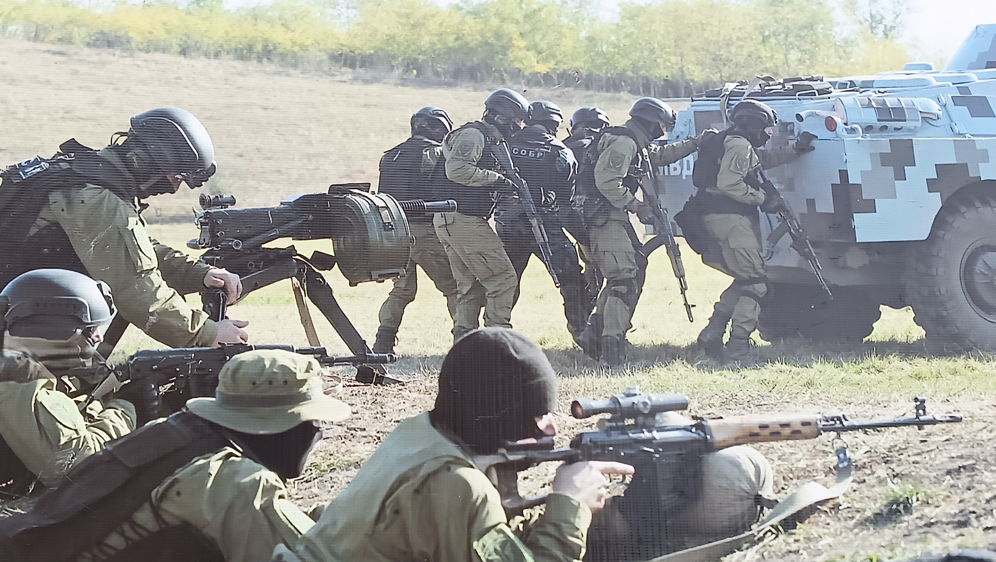 The image depicts a tactical training exercise or demonstration involving armed personnel and an armored vehicle in an outdoor, open field setting.
In the foreground, two individuals are positioned on the ground. One, wearing a balaclava and a boonie hat, is prone and aiming what appears to be a sniper rifle with a scope (likely an SVD Dragunov). Another soldier is partially visible next to him.
In the midground, a soldier in a helmet and tactical vest operates a heavy weapon, possibly an automatic grenade launcher like an AGS-17, mounted on a tripod. To the right, a group of approximately five additional soldiers, similarly equipped with helmets, tactical vests, and rifles, move in a formation or take cover. One soldier in this group clearly displays the text "СОБР" (SOBR, meaning Special Rapid Response Unit) on the back of his tactical vest.
In the background to the right, a light-colored armored personnel carrier (possibly a BTR-80 variant) with a pixelated digital camouflage pattern (grey and white tones) is visible. Two soldiers are positioned near the vehicle, one appearing to touch it while holding a rifle.
The overall scene suggests a coordinated military or law enforcement operation. The time of day is daylight, and the weather appears clear, with an open grassy field and distant trees under a bright sky. The participants are all in uniform and tactical gear, demonstrating focused engagement in their activity. The location context is Tiraspol, Moldova.