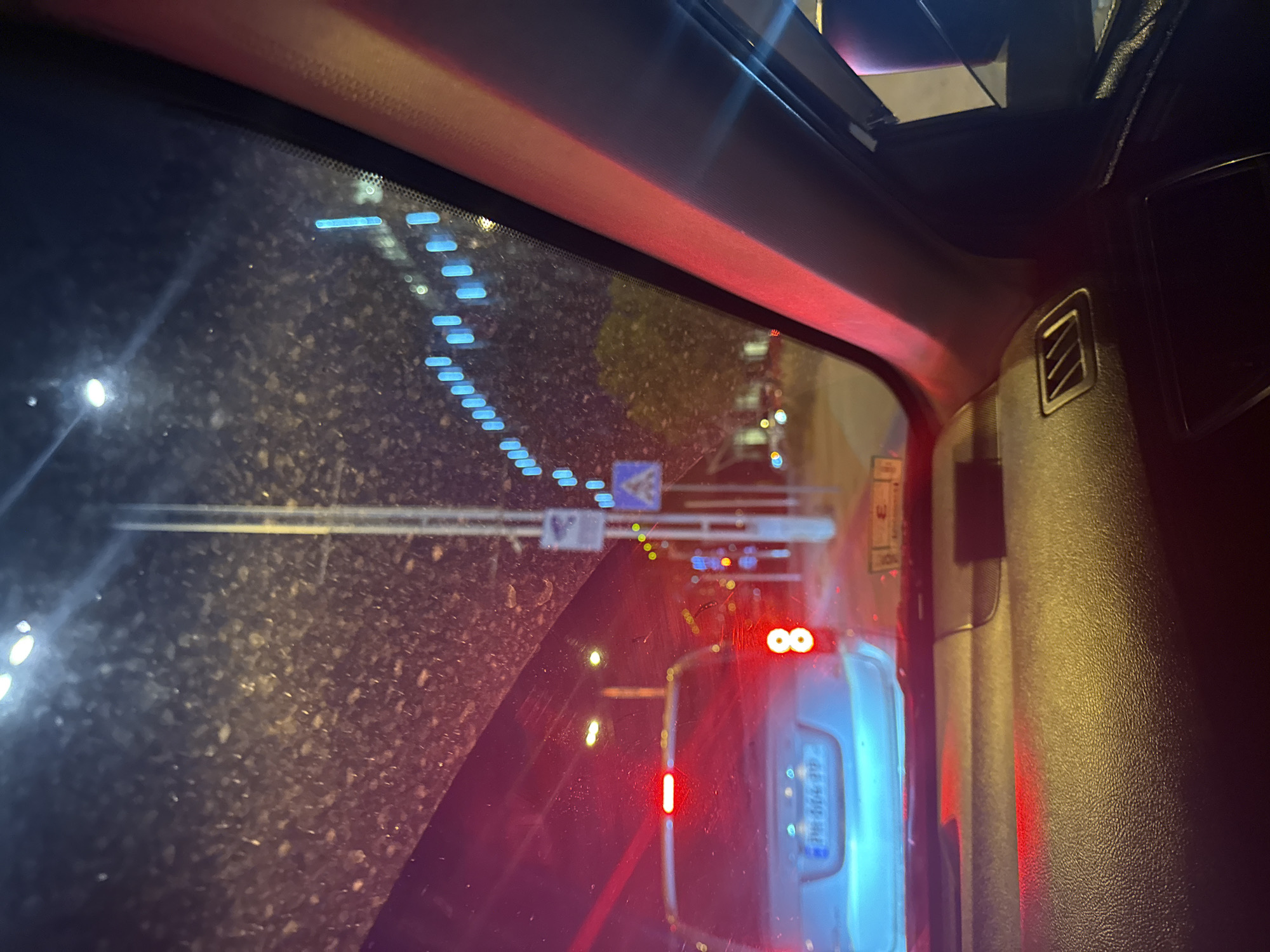 A night-time view from inside a car, looking forward through the windshield and out the side window. The windshield is smudged with dirt and shows glare from streetlights and car lights. In the foreground, the dashboard is visible, with a textured surface. A car in front has its red taillights illuminated, and a license plate is partially visible. Beyond that, streetlights cast blue and yellow light. A tall pole with signs is present on the right side of the road. The overall environment appears to be an urban setting at night.