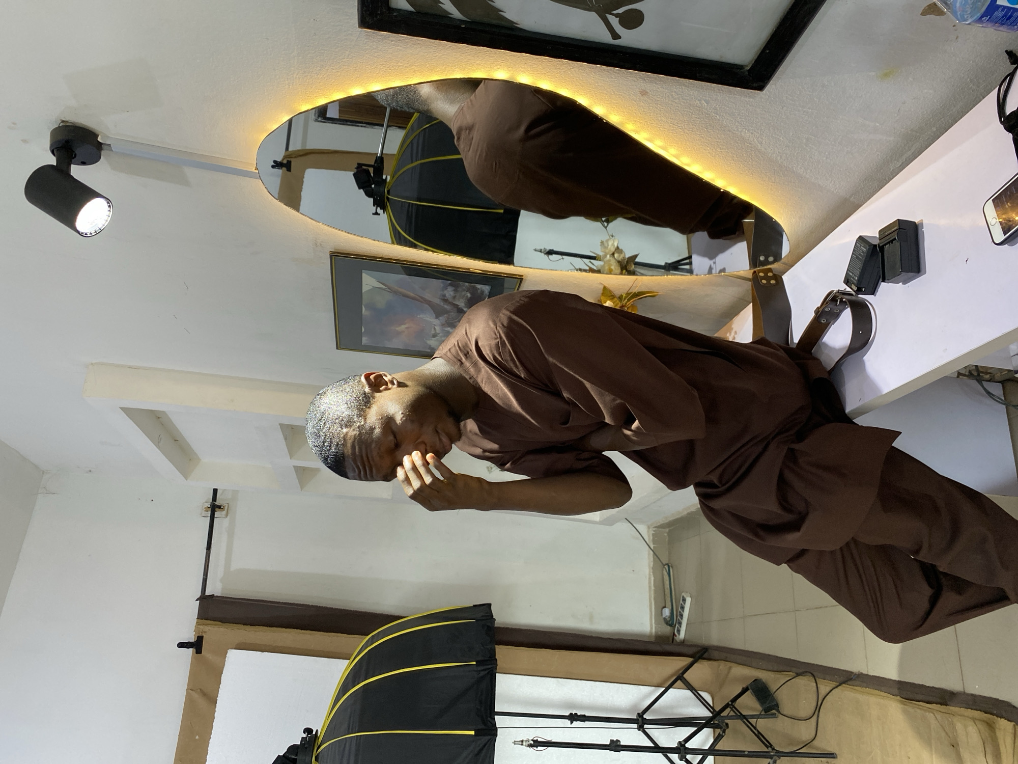 A man in a brown tunic and trousers stands inside a room, seemingly touching his forehead with his right hand. Behind him is a circular mirror with LED lights, reflecting a similar scene. To the left is a black spotlight mounted on the ceiling and a photography softbox. A table with what looks like camera batteries and a phone are on the right. A framed picture is mounted on the wall. Based on the analysis of similar images and the presence of local identifiers such as the type of softbox and clothing, the location is presumed to be in a studio or personal space in Bukuru, Nigeria.