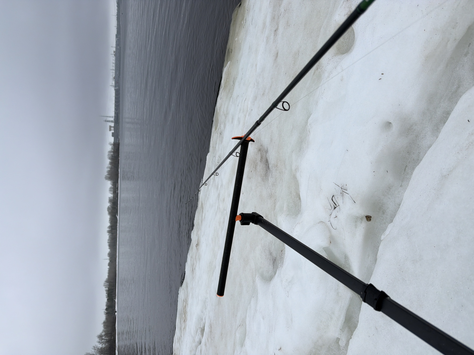 The image shows a fishing rod set up on a snowy bank, likely along the Volga River in Noviy, Russia. The rod is supported by a black stand with orange accents, and the line extends towards the water. The backdrop features a gray sky and the opposite bank of the river, with visible industrial structures and trees. The environment is wintry, as indicated by the presence of snow. There are no people visible in the scene.