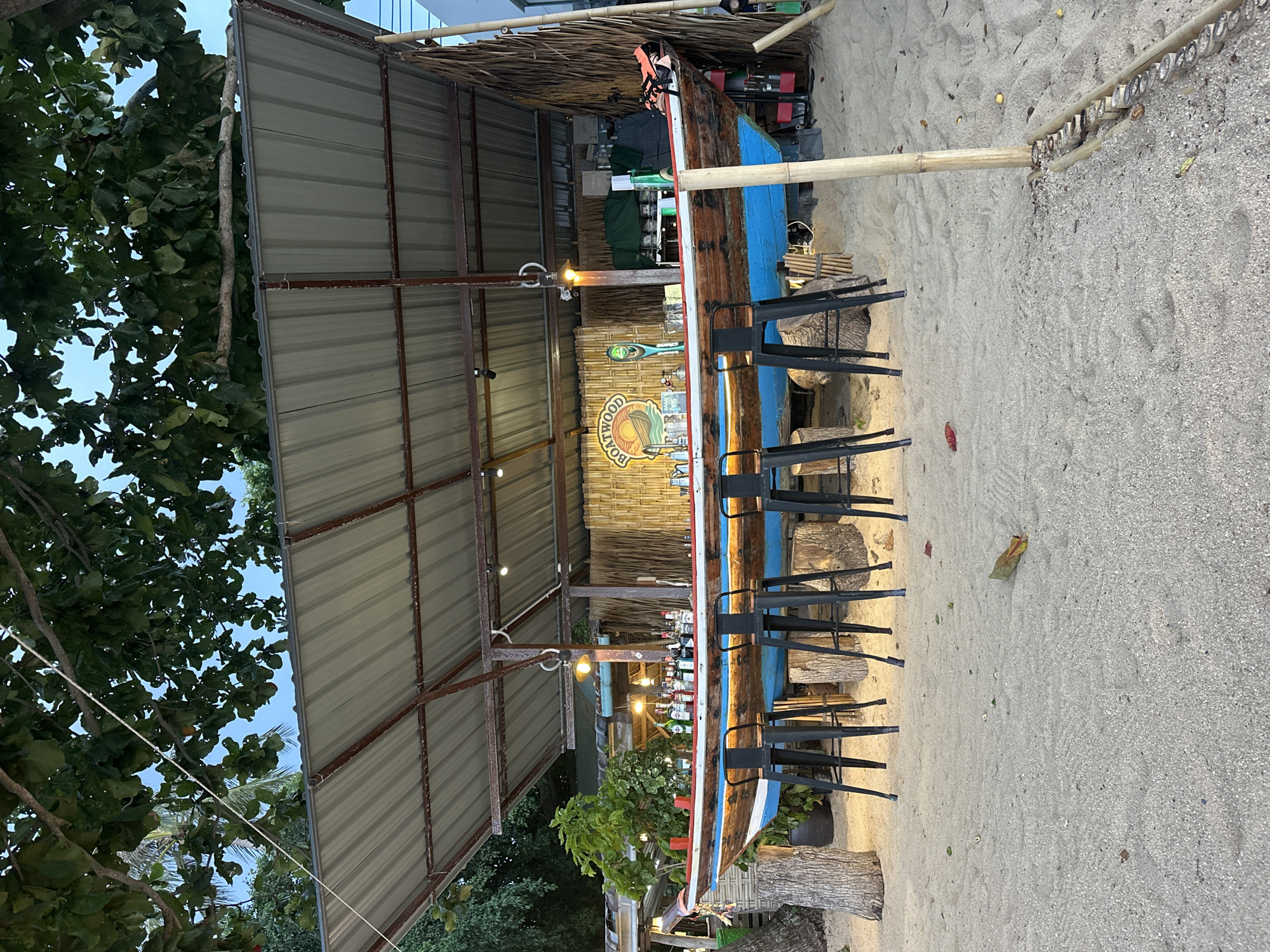 This media file depicts an outdoor bar constructed from a boat, situated on a sandy beach, likely in Baan Thong Po, Thailand. The bar, made from a weathered wooden boat with blue and white accents, is backed by a wall of woven bamboo. It is covered by a corrugated metal roof supported by metal poles and thatched roofing. Several black bar stools are arranged in front of the boat bar, with a few tree stumps also serving as rustic seating or decorative elements on the sand. Behind the bar, rows of bottles are visible, suggesting it is stocked and operational. The scene is illuminated by a few warm-toned lights, indicating it may be late afternoon or early evening. Lush green trees with large leaves partially frame the left side of the scene, and the sand is scattered with small debris and footprints. The overall atmosphere appears relaxed and tropical, suitable for a beachside establishment. A circular logo with text is visible on the bamboo wall behind the bar. The text on the logo reads "BOATWOOD" in a rustic font, with imagery of a palm tree and a wave.