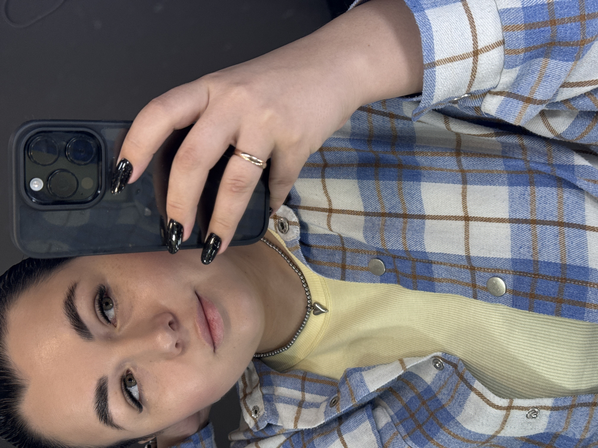 A young woman is taking a selfie. She has dark hair, light skin, and wears dark mascara. Her fingernails are painted black with glitter. She is holding a black cell phone in her right hand, on which she wears a gold ring. She is wearing a light yellow t-shirt under a blue and white plaid shirt with brown accents and silver buttons. She has a silver chain necklace with a small heart pendant. It is not possible to confirm if the photo was taken in Chaikovsky, Russia.