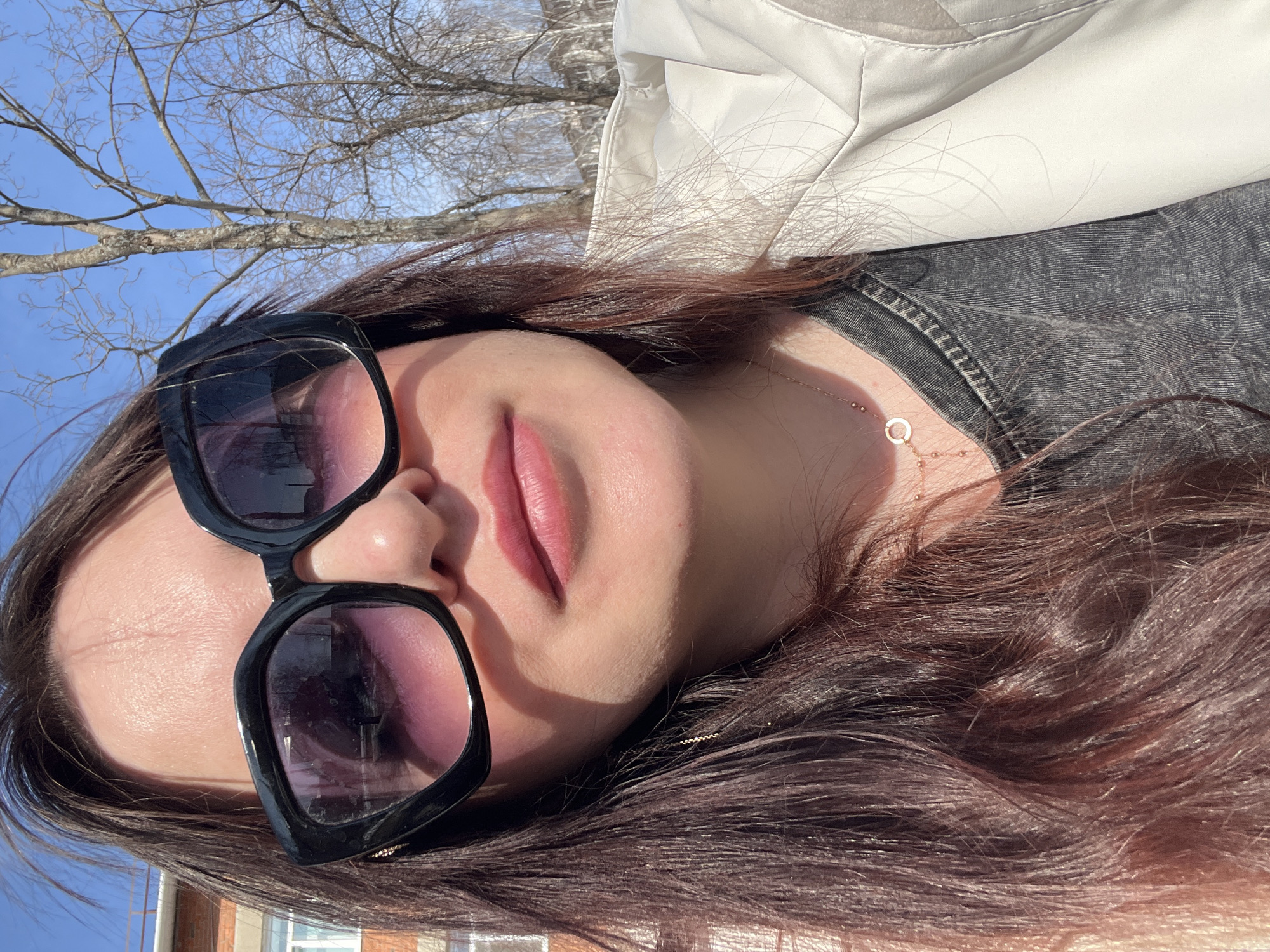 A photograph shows a fair-skinned woman wearing large, black-rimmed sunglasses with a purple tint, a gray denim shirt, and a cream-colored quilted jacket. She has dark brown hair and is wearing a delicate gold necklace. The woman is outdoors, with bare tree branches visible in the background against a blue sky. A portion of a brick building with a white-framed window is visible in the lower left corner. The photograph appears to have been taken on a sunny day. No definitive evidence allows for a determination of the location in the city of Noviy, Russia.