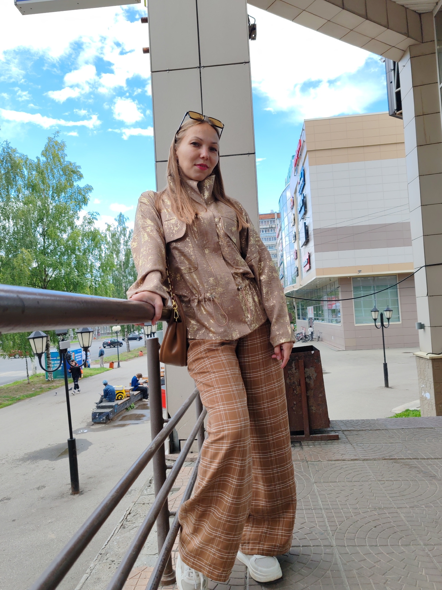 A full-body outdoor shot shows a young woman standing on a balcony, leaning on a railing. She is wearing a bronze jacket with gold accents and matching plaid pants, white sneakers, and sunglasses on her head. A brown purse is slung over her shoulder. Behind her, a modern building with a beige and white facade dominates the background, with trees and a cloudy sky above. In the distance, people are seen on a street with cars and lampposts. The scene appears to be a commercial district in Syktyvkar, Russia.
