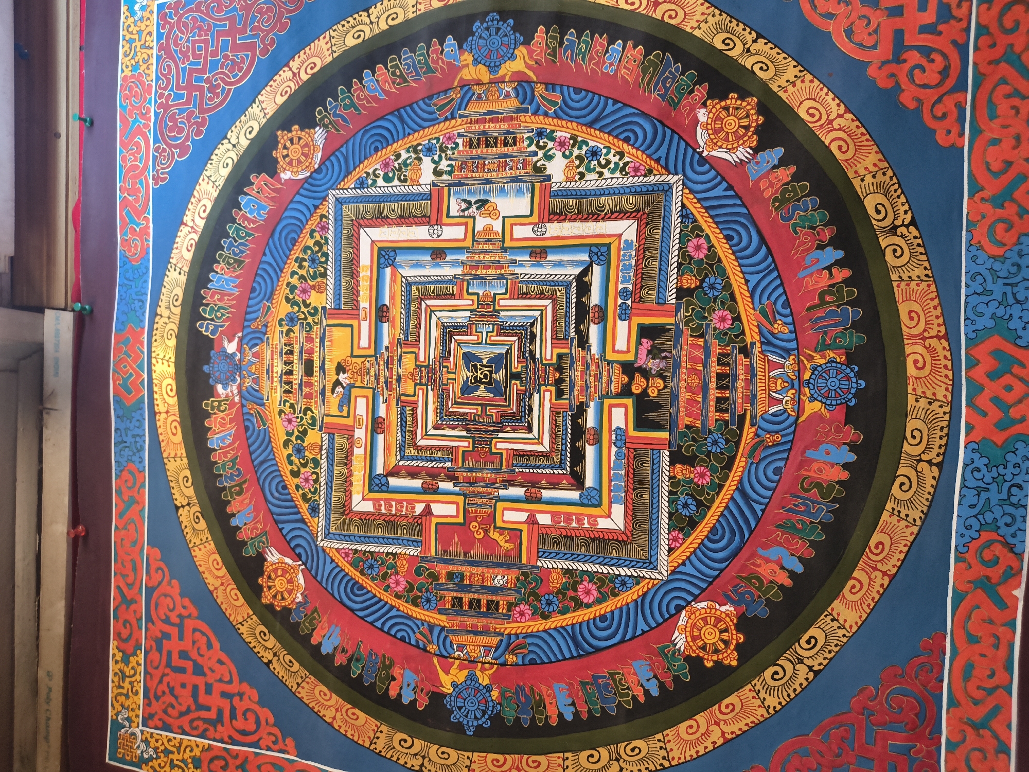 The image shows a vibrantly colored Tibetan mandala painting hanging on a wall. The mandala is a complex geometric design with intricate patterns and symbolic imagery, featuring concentric squares and circles, lotus flowers, dharmachakras (wheels of dharma), and stylized figures. The colors are rich and varied, including blues, reds, yellows, greens, and gold. The painting appears to be made of fabric or canvas and is bordered by a dark red fabric. The top of the image shows a portion of a wooden structure and a dark red border, suggesting the painting is displayed indoors, possibly in a shop or a private residence. The painting is likely a thangka, a traditional Tibetan scroll painting. There are no people or actions depicted in the image. The location is identified as Bhagsu, India, implying this might be a piece of art found in the area known for its Tibetan culture and tourism.