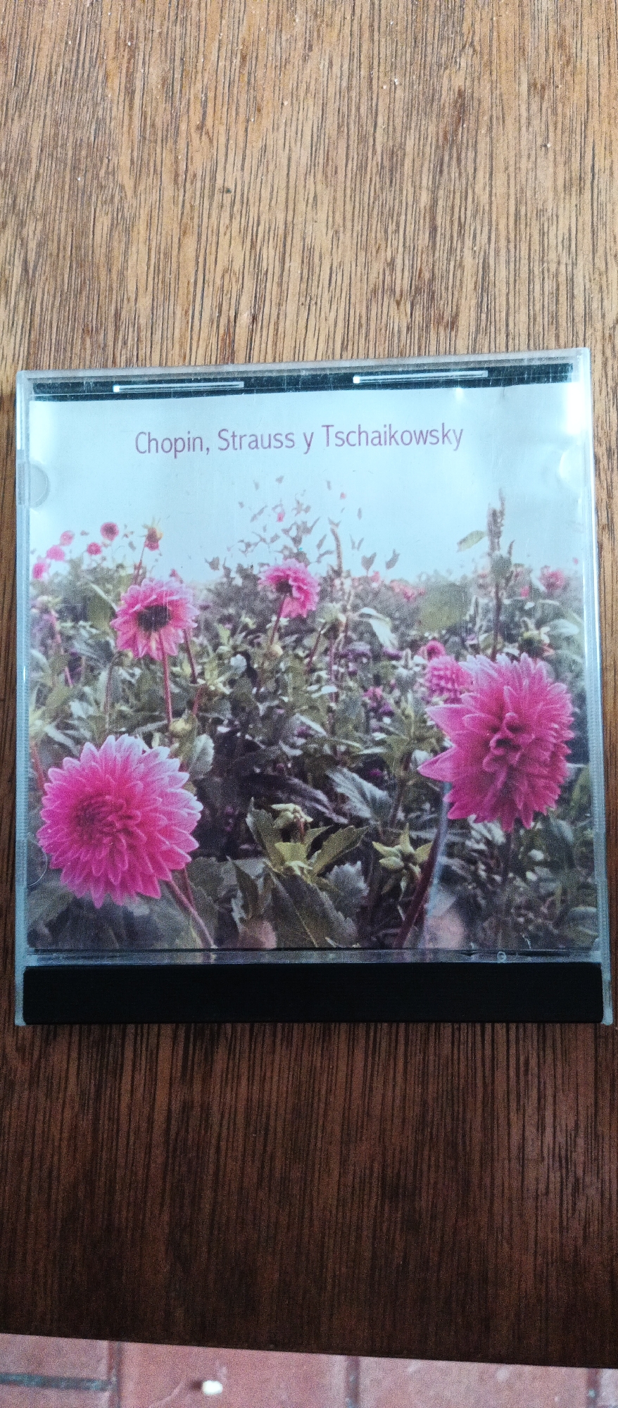 This is a close-up, overhead shot of a CD case lying on a wooden surface. The CD case is open, revealing a CD with a cover image of a field of pink dahlias. The text on the cover reads "Chopin, Strauss y Tschaikowsky" in a purple font. The wooden surface appears to be a table or shelf, with visible wood grain and some dust particles. The background also shows a hint of red brick. The lighting is diffused, suggesting it could be daytime, but no specific time can be determined. The overall mood is calm and serene, evoked by the floral imagery.