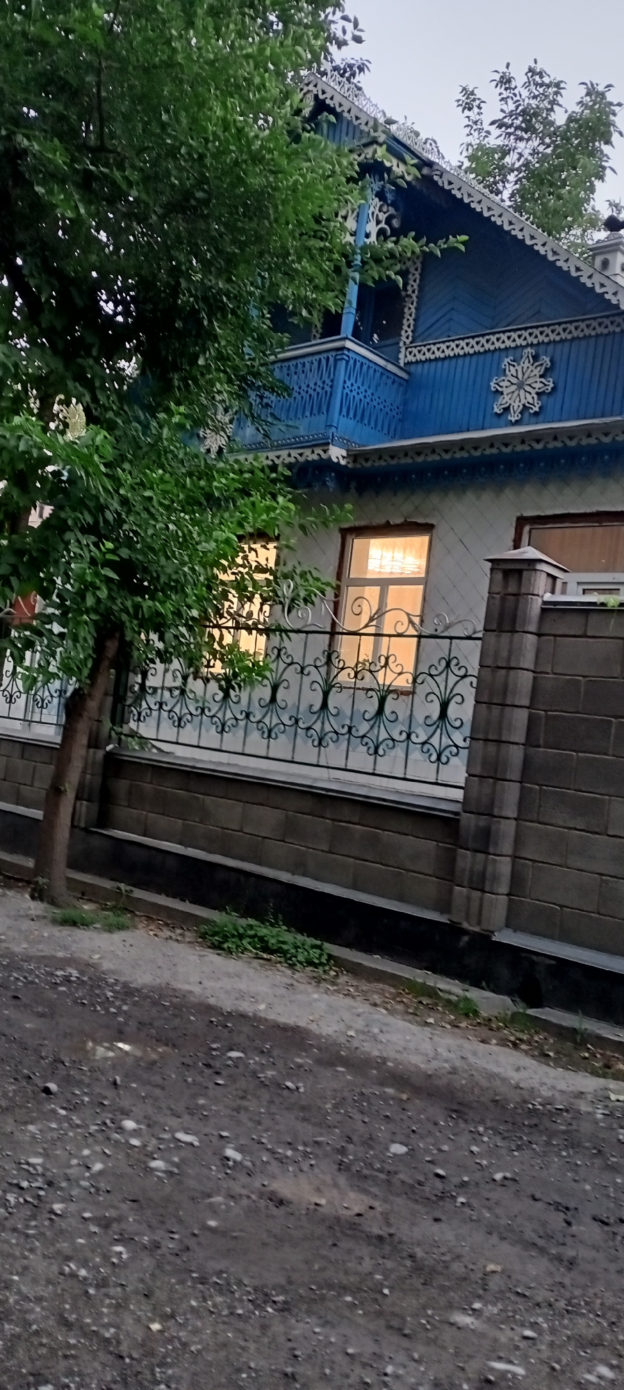 In Romanovka, Kyrgyzstan, a traditional blue wooden house with ornate white trim and a balcony is viewed from a dirt road. A large green tree partially obscures the house. A decorative metal fence runs along the property line, with a grey brick wall beneath it. The windows of the house are lit from within, casting a warm glow on the exterior. The scene appears to be set during dusk, with the sky a pale grey.