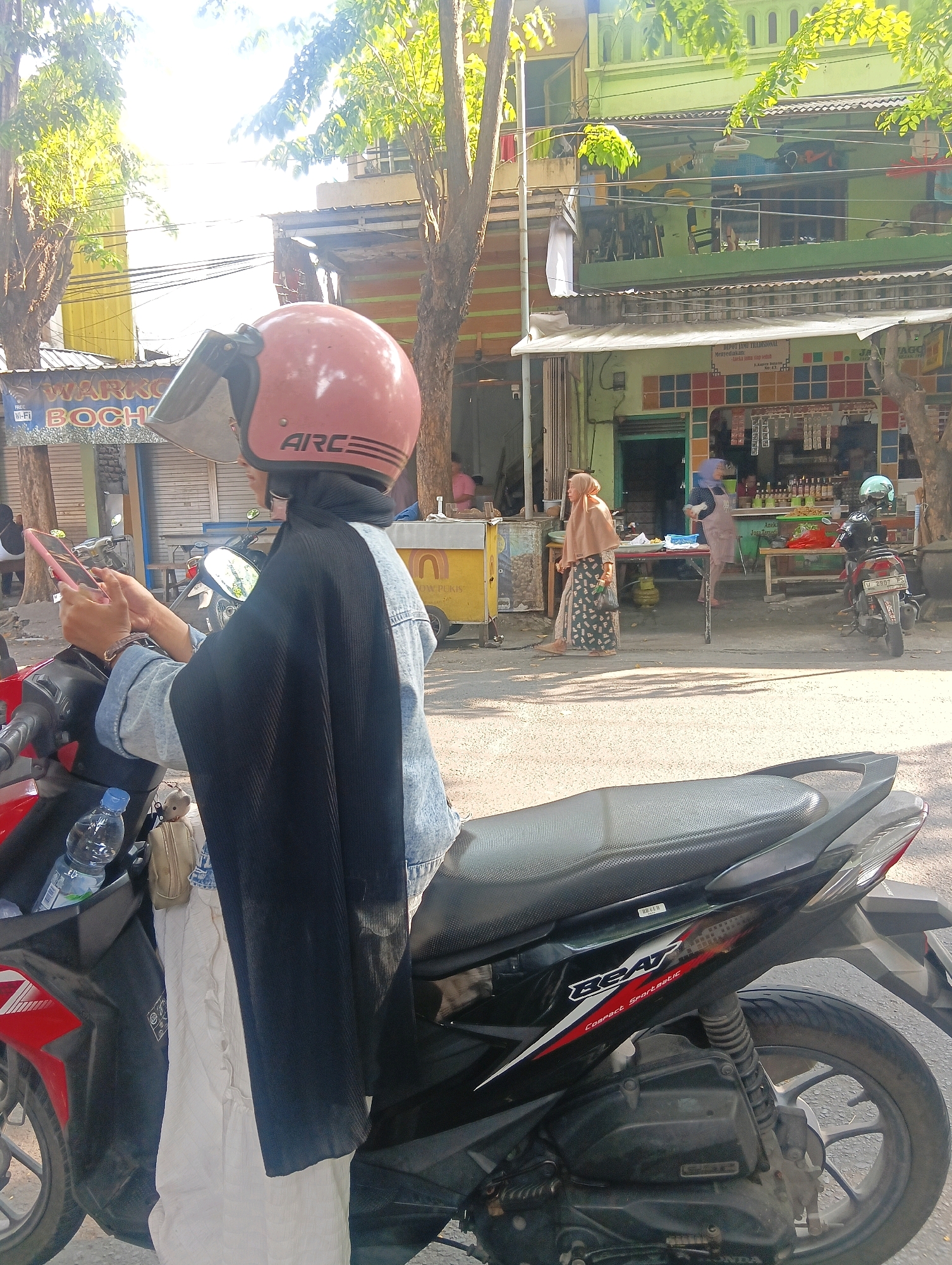 In Gapurosukolilo, Indonesia, a person wearing a light blue denim jacket, a black headscarf, and a black outer garment sits on a black and red motorcycle. The person is holding a pink smartphone in their hands and looking at it. On the motorcycle's rear seat, a bottle of water and a small teddy bear are visible. In the background, several buildings with green and yellow facades line the street, with trees casting shadows. Two other individuals are visible further down the street, one walking and the other standing near a stall. Red and white signage is present on some of the buildings.