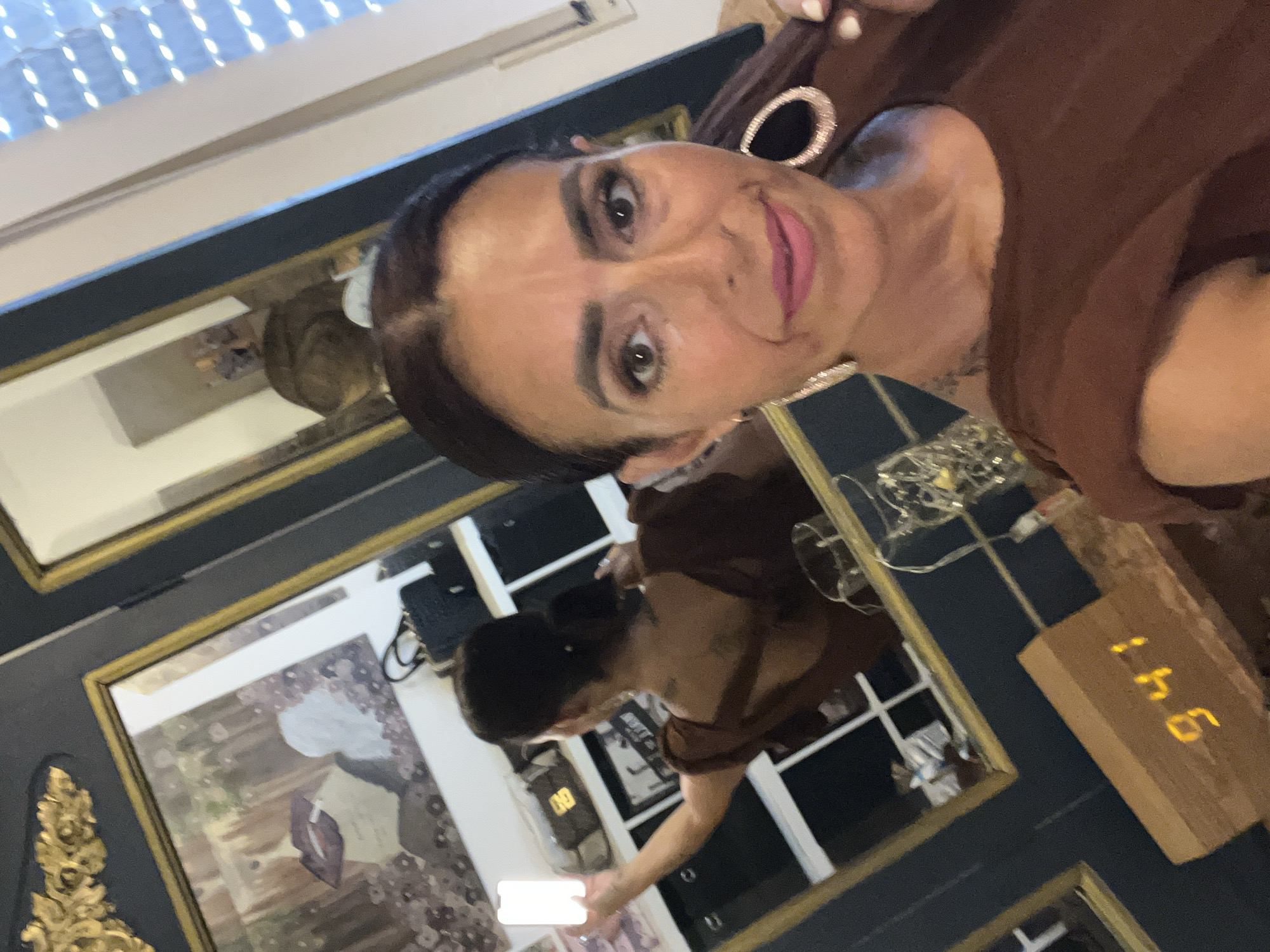 The image is a selfie taken indoors, possibly in an apartment, with a mirror reflecting some of the environment. The main subject is a woman with dark hair and makeup, wearing a brown top and large hoop earrings. She is holding a white smartphone, which is illuminated. In the background, a mirror shows her reflection from behind, revealing tattoos on her back and a low-cut top. Various objects are visible in the reflection, including shelves with handbags and decorative items. A digital clock displaying "9:41" is visible in the foreground on a wooden surface. The scene appears to be in Konz, Germany.