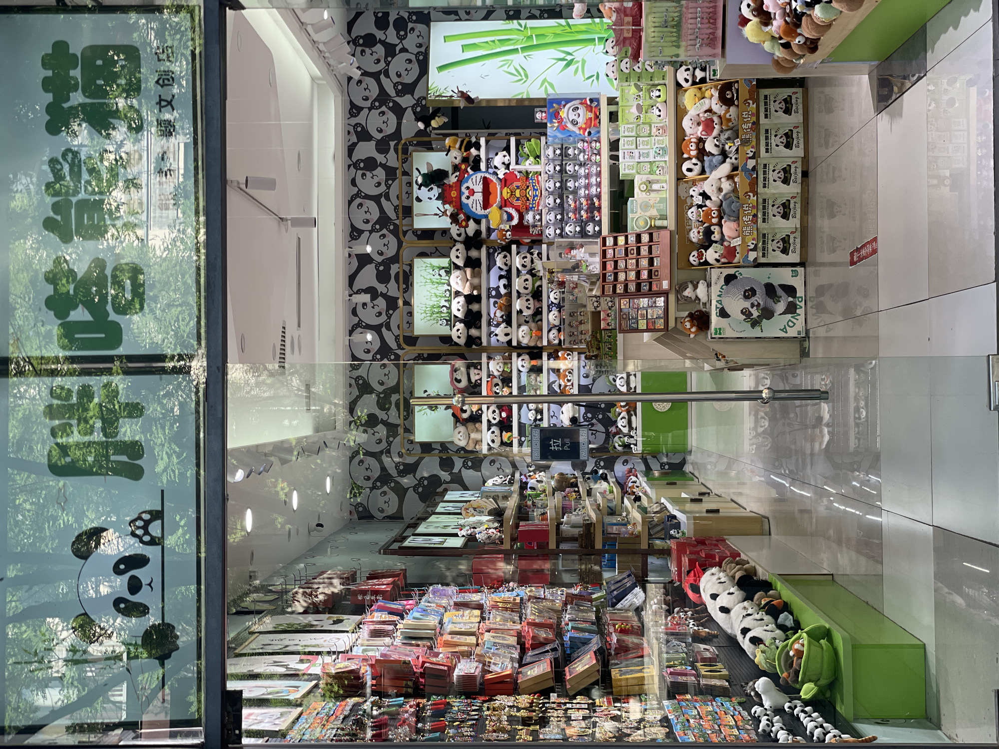 The image captures the interior of a retail store, possibly a souvenir shop, located in Dongcheng District, China, specialized in panda-themed merchandise. The store's interior is visible through a glass storefront with reflections of the outside environment. Inside, shelves are filled with panda plush toys of varying sizes, along with framed artwork, trinkets, and stationery featuring pandas. The wallpaper consists of a repeating pattern of panda faces. A sign on the storefront includes Chinese characters, including "胖嗒熊猫", which means "Fat Panda" and 李顯文创店 which means "Li Xian Cultural and Creative Store". This is supported by the various pieces of panda-themed merchandise being offered. The overall impression is that of a store that caters to tourists or locals seeking panda-themed souvenirs or gifts.