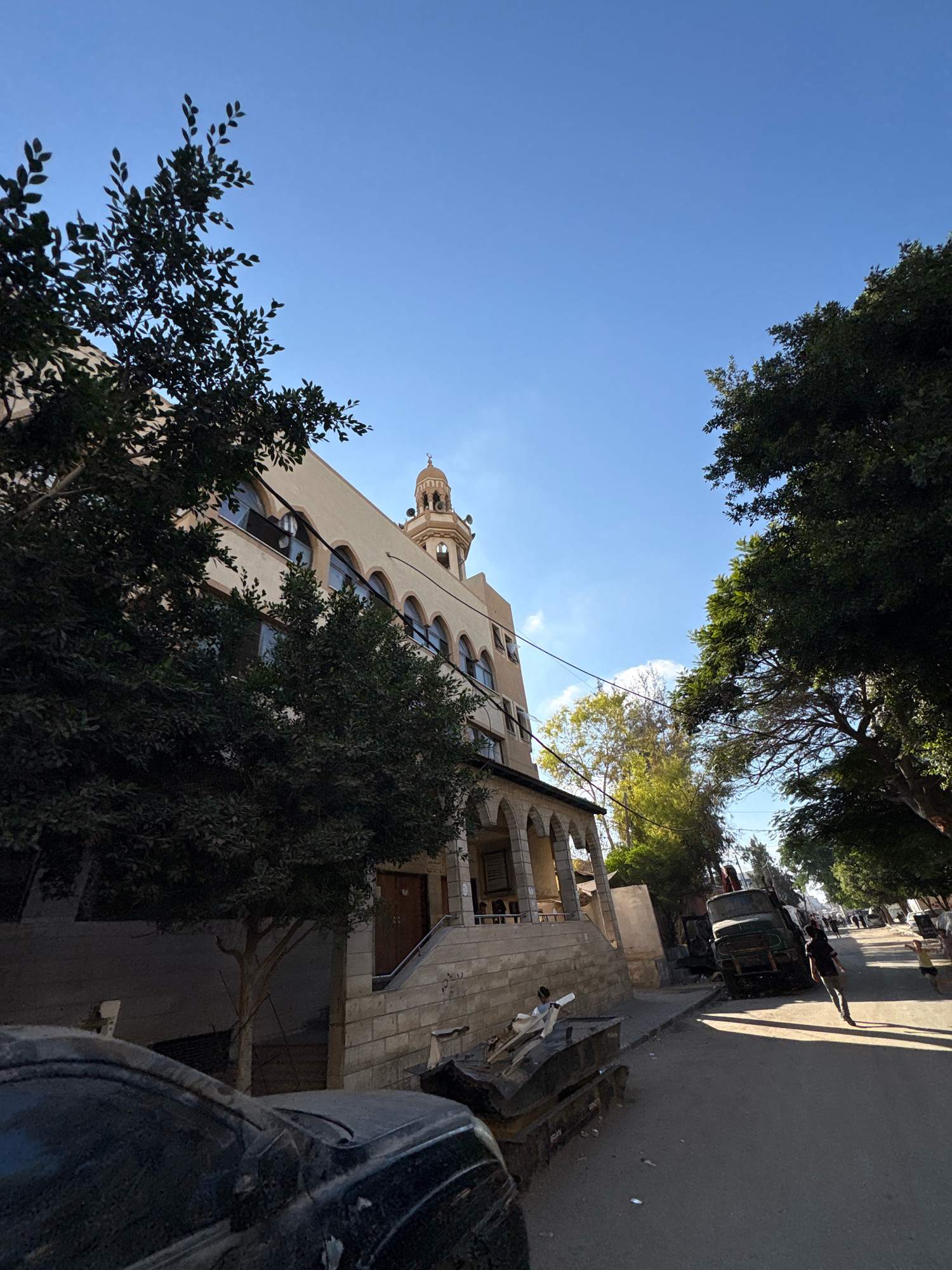 The image depicts a street scene in Gaza City, Palestinian Territories. On the left, a multi-story building with arched windows and a prominent minaret of a mosque is visible, partially obscured by trees. In front of the building, there are stone steps leading up to an entrance. The street itself is paved, with several vehicles present. A dark-colored car is parked in the foreground. Further down the street, a green truck is parked at an angle, and a person is walking away from the camera. Several other people are visible in the distance, and trees line the right side of the street. The sky is clear and blue, indicating daytime. The overall impression is of a street in an urban environment, possibly with some signs of disrepair or recent activity, given the presence of what appears to be debris in the street.