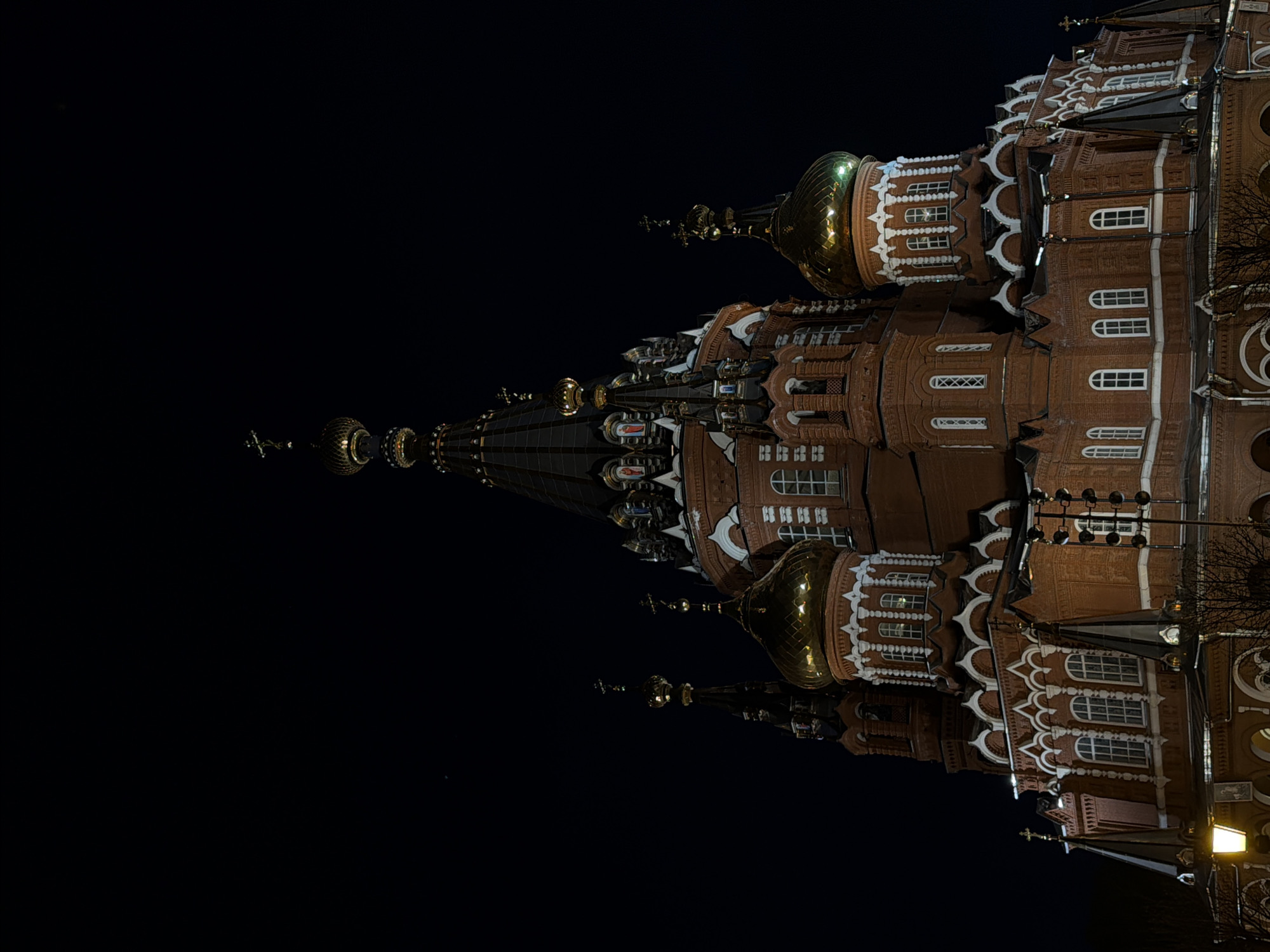 The image shows the exterior of St. Michael's Cathedral in Izhevsk, Russia, at night. The cathedral, made of red brick with white trim, has multiple golden domes and spires. The sky is dark, suggesting nighttime, and some details of the building are illuminated by artificial light.