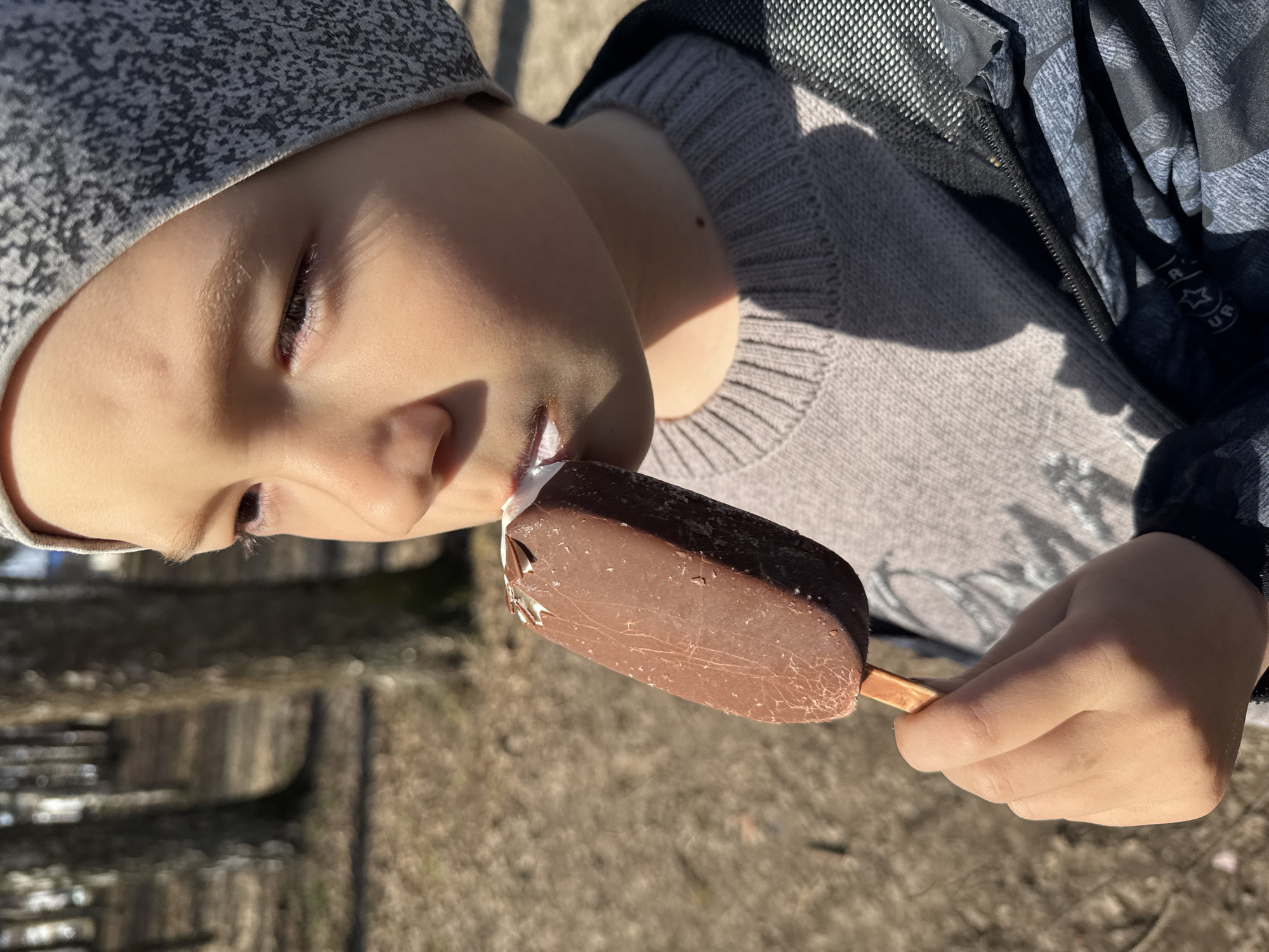 A child wearing a grey beanie and a grey sweater is eating a chocolate-covered ice cream bar. The child is holding the wooden stick of the ice cream bar with their right hand. The location is Dmitrov, Russia. The background consists of a blurred outdoor setting that appears to be a park or similar area.