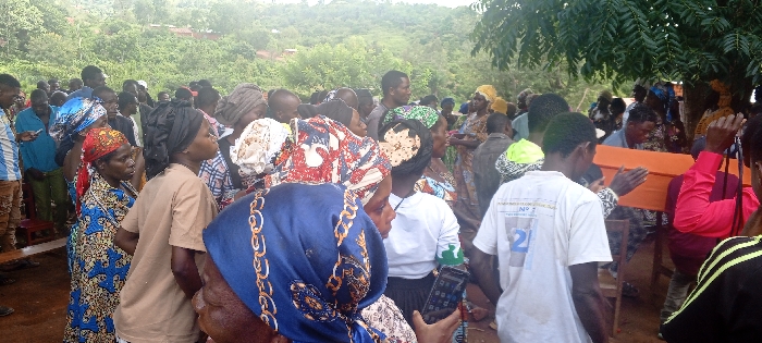 A large crowd gathers in an outdoor setting, seemingly a village. Many people wear head coverings or hats. An orange coffin is visible, carried by two individuals. The crowd seems to be gathered around it. The scene takes place on a day with overcast conditions, with some trees and greenery visible in the background. Some individuals hold or operate mobile phones.