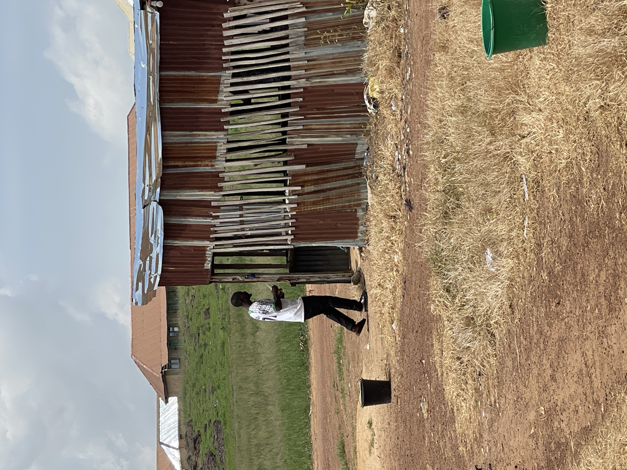 In Durumi, Nigeria, a lone individual walks past a dilapidated corrugated metal structure. The structure, appearing to be a makeshift stall or shelter, has a rusted, uneven corrugated metal roof and walls composed of vertical, weathered wooden slats. In front of the structure, a black bucket stands on the dirt ground. The person, wearing a white t-shirt with a green number on the back and black pants, is walking from left to right, holding a white object, possibly a phone, in their right hand. Behind the stall, a grassy hill is visible, with several buildings featuring brown and silver roofs in the distance. The foreground is a dry, dusty terrain with patches of dead grass and scattered debris. A bright green bucket is situated in the dry grass to the right of the frame. The sky is partly cloudy.