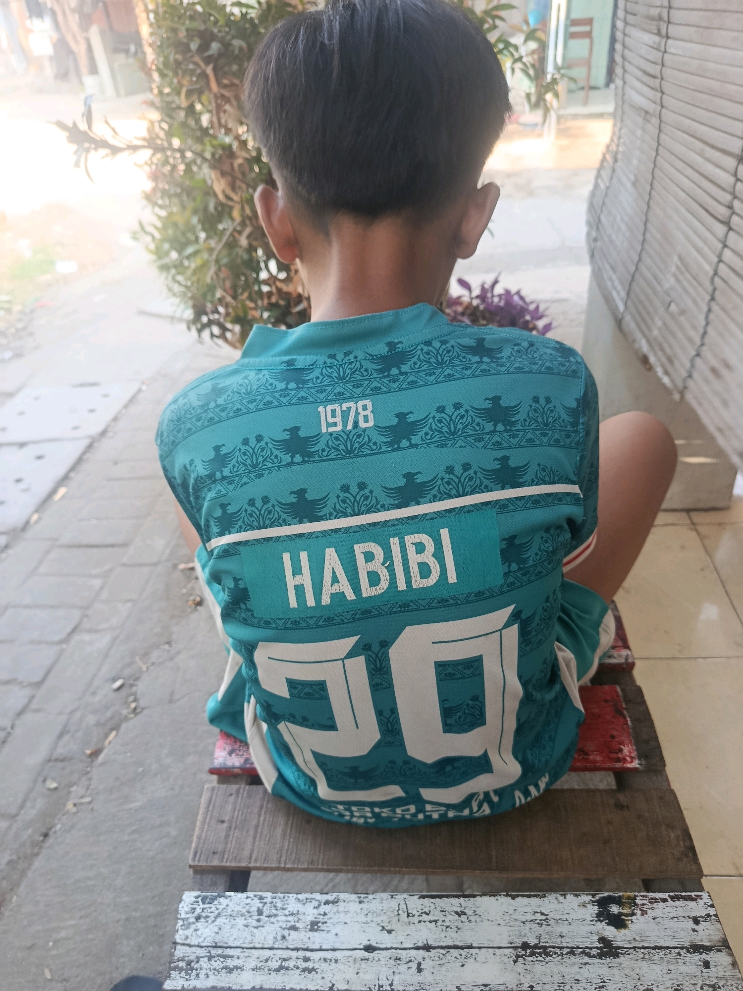 A rear view of a young boy wearing a teal-colored t-shirt sits on a wooden bench. The t-shirt has a repeating pattern of eagle-like figures and decorative elements. The number "1978" is printed on the upper back, above the word "HABIBI" in large white letters. Below "HABIBI", a large white number "29" is visible. The boy is positioned outside, with a blurred background of plants, a wall made of woven material, and paved ground. The lighting suggests it is daytime, possibly outdoors in a residential area of Jobokuto, Indonesia.