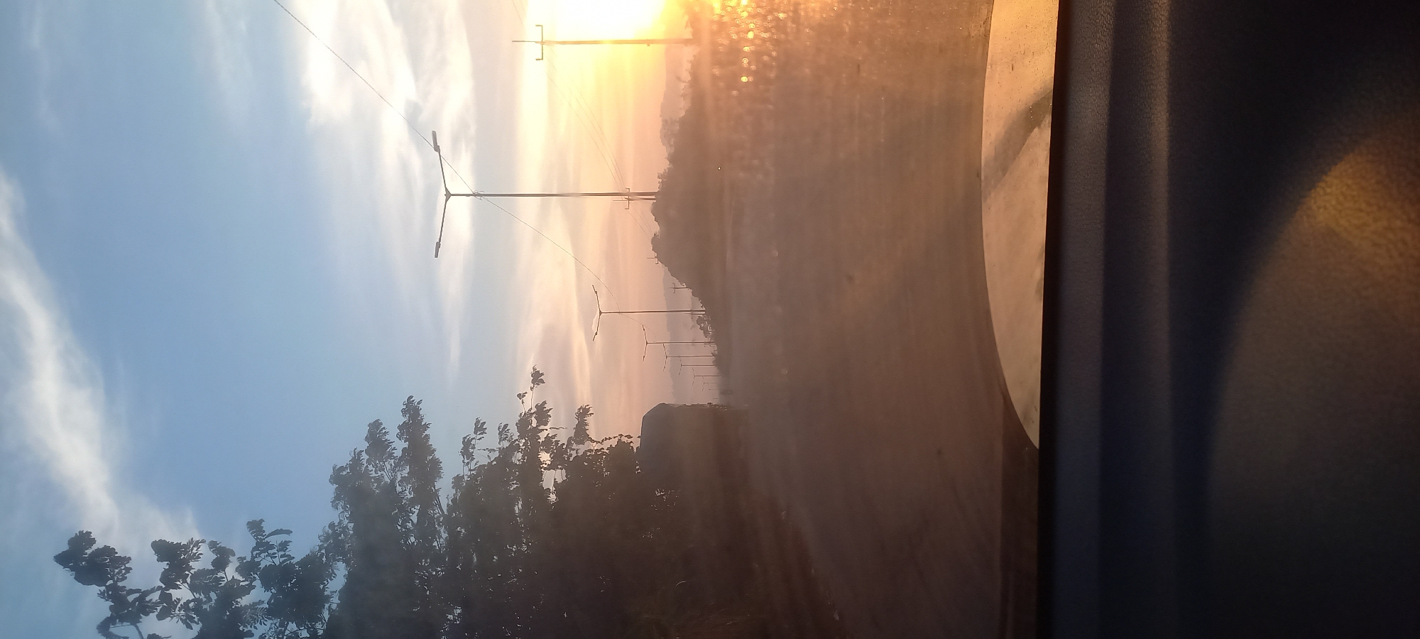 The image captures a roadside scene at sunrise in Sepakat, Indonesia. A road stretches into the distance, lined with streetlights. The sky is a gradient of blue and orange, with some wispy clouds. The sun is low on the horizon, casting a warm glow over the landscape and creating reflections on the road, possibly due to moisture or dirt. A dense cluster of trees is visible on the left side of the frame. The foreground shows the interior of a vehicle, with the dashboard and windshield partially visible. The overall atmosphere is serene, suggesting an early morning commute or journey.