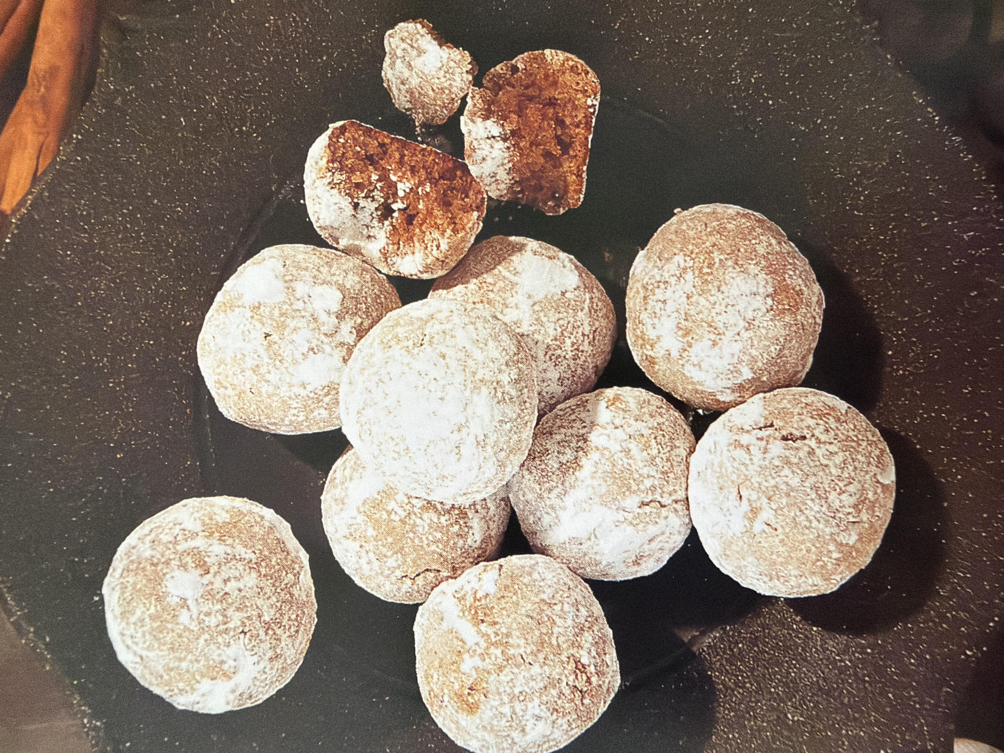 A close-up, overhead shot shows a collection of round, powdery cookies scattered across a dark, textured surface, likely a plate or serving dish. The cookies are a warm brown color and coated with a generous dusting of white powdered sugar, giving them a rustic, homemade appearance. Several cookies are whole, while a few are broken, revealing a dense, crumbly interior. The background is dark and speckled, with a hint of cinnamon sticks visible in the upper right corner. The lighting creates soft shadows, emphasizing the three-dimensional texture of the cookies. No people or city elements are present in the image.
