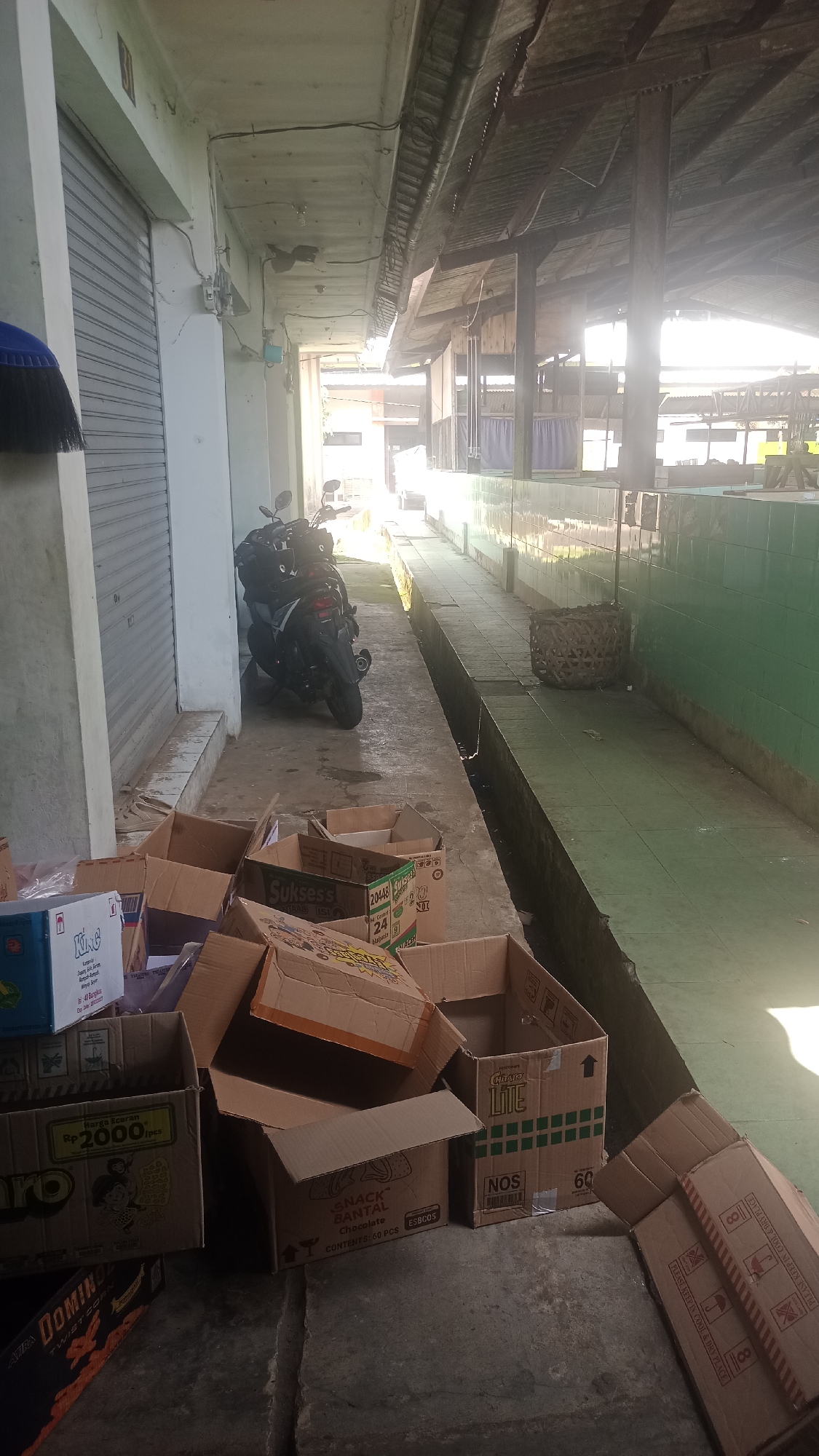 A motorcycle is parked next to a closed metal roll-up door, with several cardboard boxes piled on the ground in the foreground. The boxes are of various sizes and some are open. The scene appears to be outdoors, possibly in a market or alleyway, with a covered structure in the background and a tiled green wall on the right. The lighting suggests it is daytime, with sunlight casting shadows. The ground is concrete with visible cracks. A blue-bristled broom is partially visible to the left of the motorcycle. The overall impression is one of a storage or delivery area.