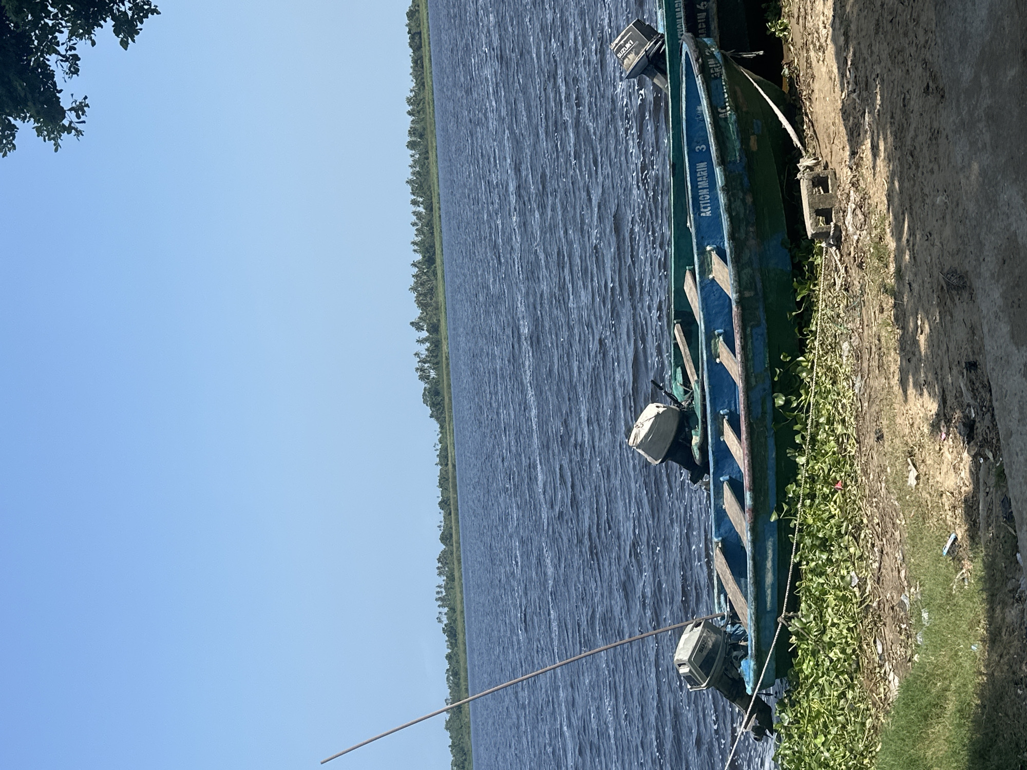 The image depicts two small motorboats pulled ashore on a sandy embankment along a body of water. The boats are green and blue with wooden seats and outboard motors. The water is dark blue and choppy, with a line of lush green vegetation on the distant shore, including palm trees. The sky is a clear, light blue. The embankment in the foreground is sandy and littered with debris, including green aquatic plants. A tall, thin pole rises from the water near the left boat. The scene appears to be a waterfront area in Badagry, Nigeria, likely a fishing or transportation hub.
