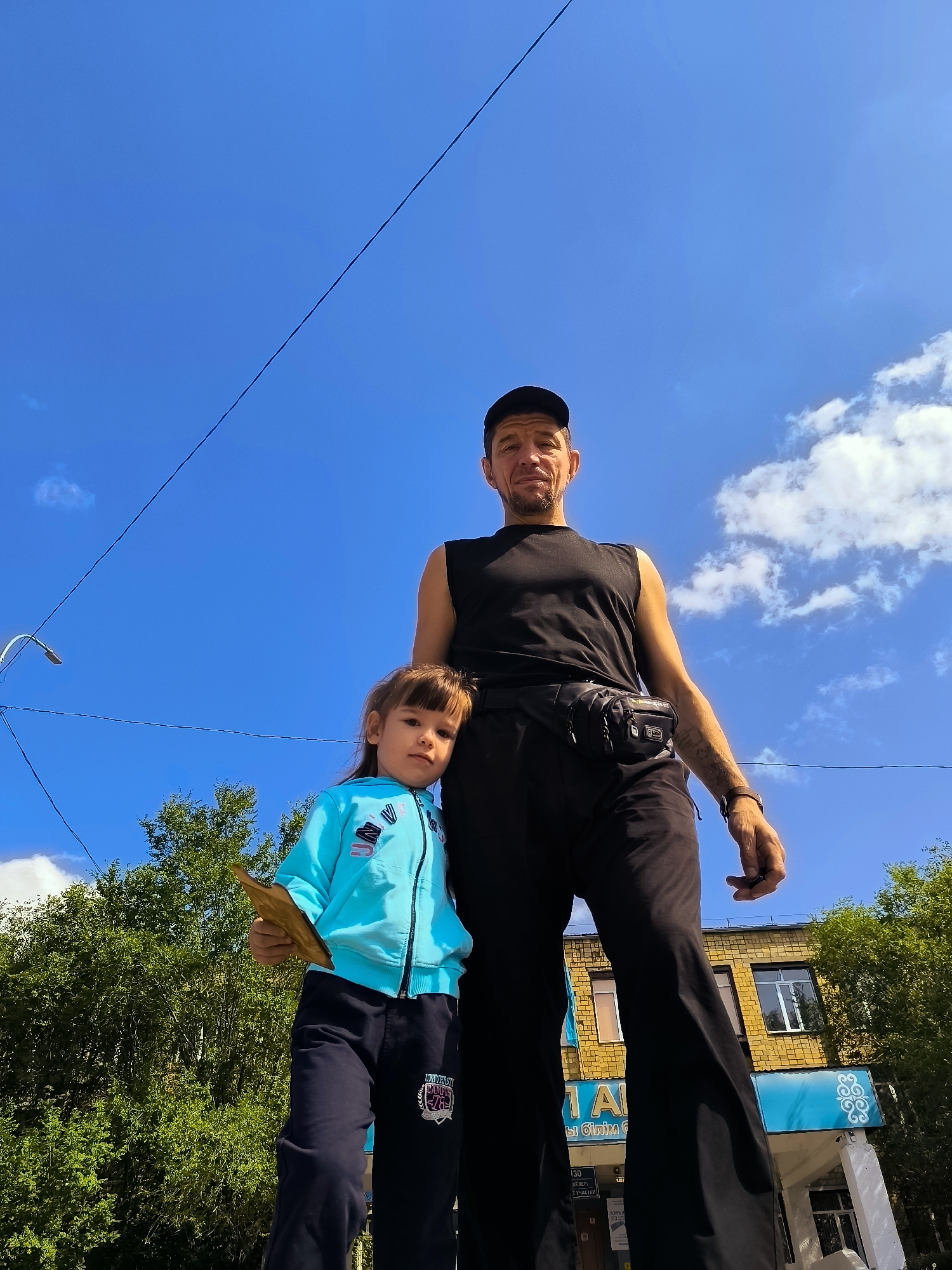 A low-angle, full-body shot captures a man and a young girl standing outdoors under a bright blue sky with scattered clouds. The man, wearing a black sleeveless shirt, black baseball cap, and black pants with a fanny pack, stands slightly behind and to the right of the girl. He has short facial hair and tattoos are visible on his right arm. The girl, wearing a light blue hooded jacket and dark blue track pants, stands in front of the man, facing the camera. She is holding a light-colored, flat object in her left hand.
In the background, a yellow brick building with windows and a light blue sign with white text and a decorative emblem is visible. Green trees flank the building on the left and right sides. A street light on a pole is positioned on the far left of the frame, and power lines stretch across the sky. The scene appears to be an urban environment, possibly in Qarağandı city administration, Kazakhstan.