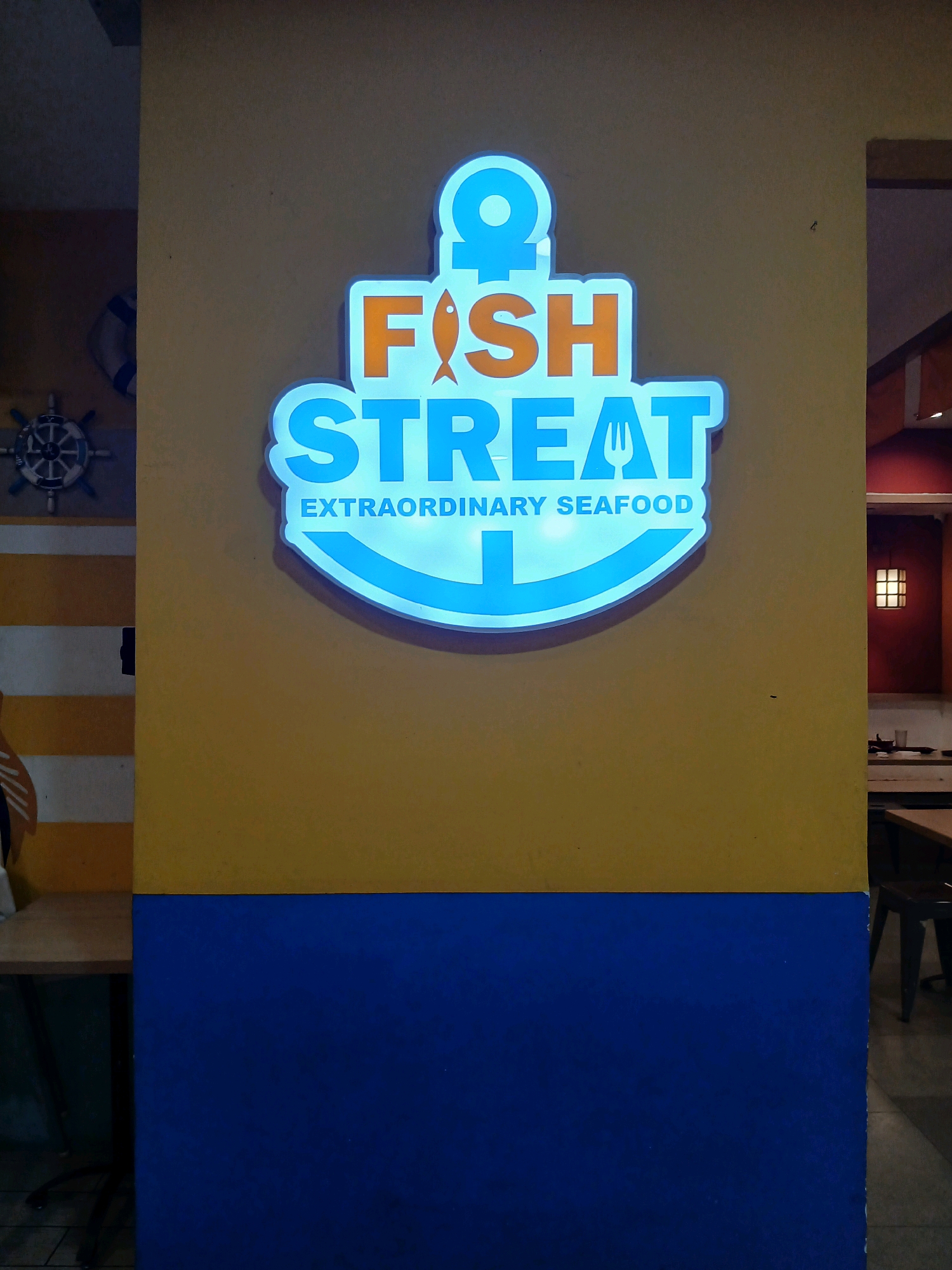 A neon sign shaped like an anchor reads "FISH STREAT" in orange and blue letters, with "EXTRAORDINARY SEAFOOD" written in smaller blue letters below. The sign is mounted on a yellow wall. To the left of the sign, a ship's wheel and a life preserver are visible, suggesting a nautical theme. Below the yellow wall is a blue section. In the background, a restaurant interior is visible with tables and chairs, and a decorative lantern hangs on a red wall. The location is identified as South Jakarta, Indonesia. There are no people visible in the image.
