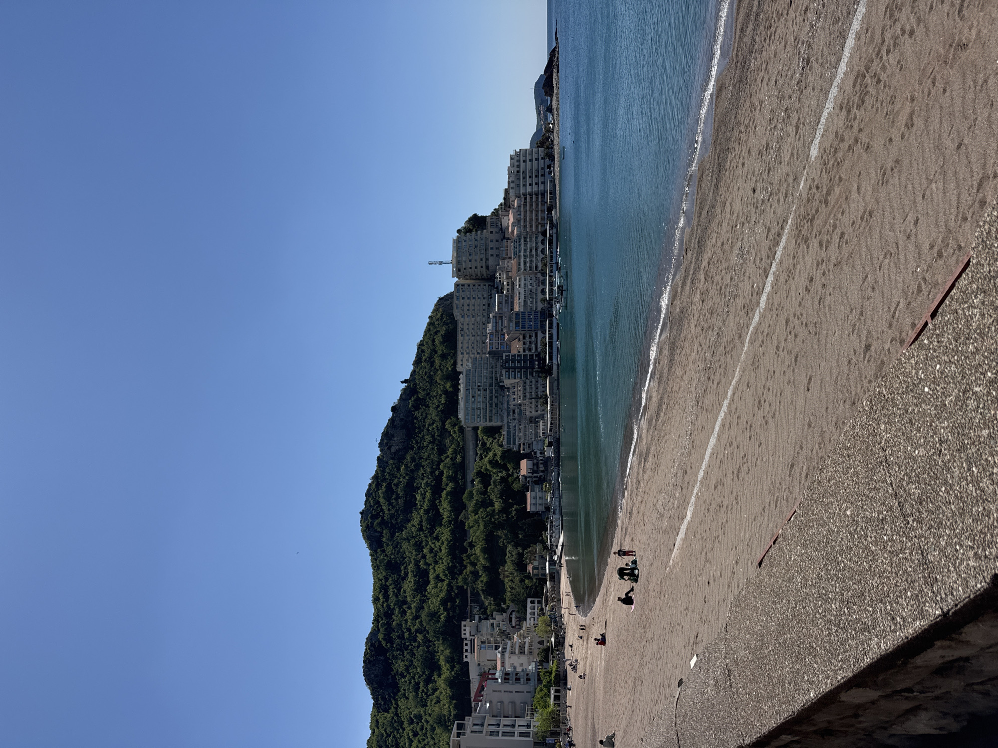 The image depicts a view of Rafailovići, Montenegro, a coastal town, likely taken in daylight under clear, blue skies. The left side shows a steep hillside densely covered with green trees, blending into a cityscape consisting of multi-story buildings and houses layered into the terrain. The right side reveals a beach with dark sand and a lighter strip bordering the water. The Adriatic sea is visible, appearing calm and blue-green. On the beach, several small groups of people are scattered around. The very bottom-right of the frame shows a rough, concrete surface, indicating the vantage point is likely from a pier or elevated walkway.
