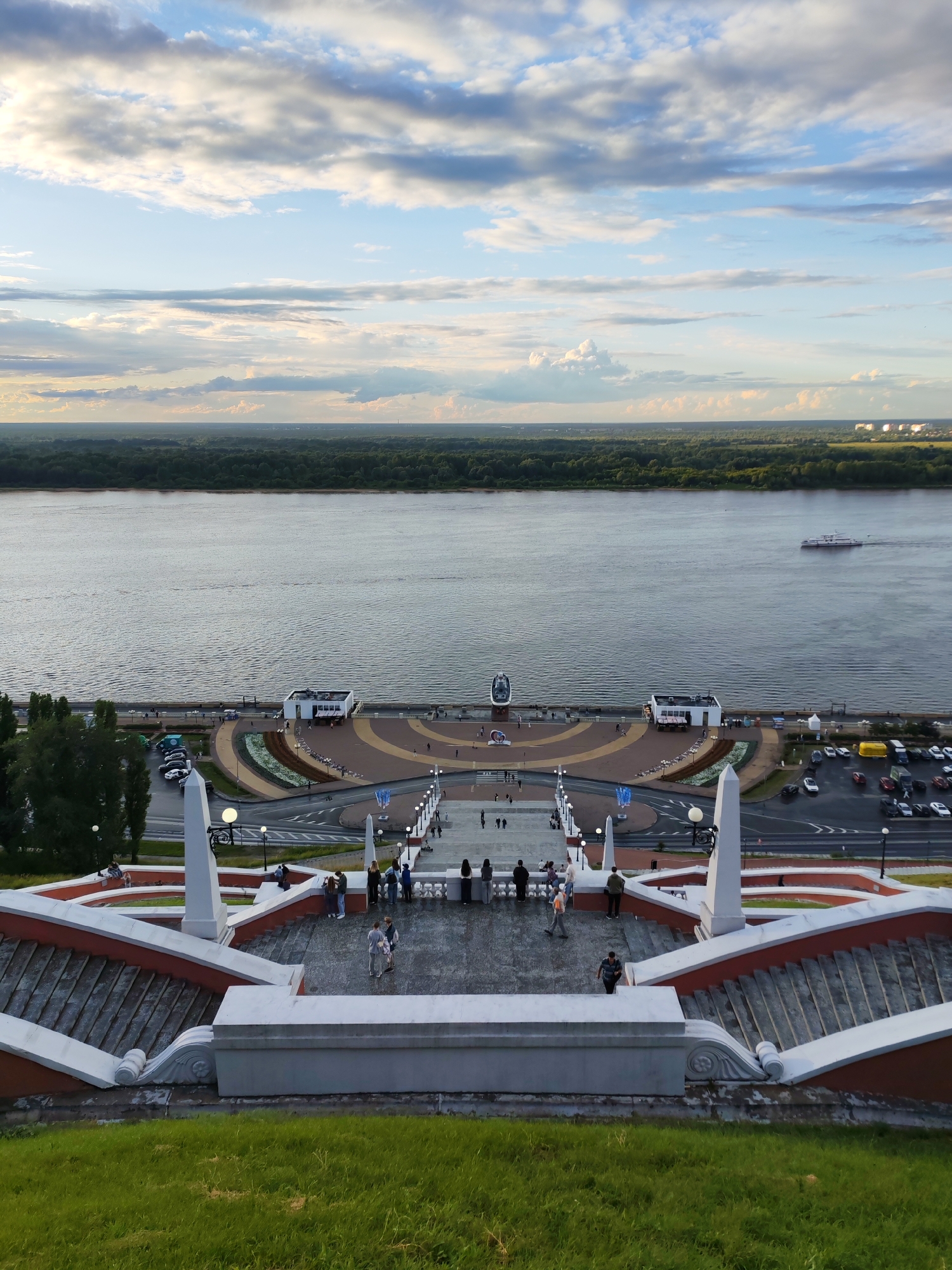 The image depicts a scenic view from the Nizhny Novgorod Kremlin's embankment, overlooking the confluence of the Volga and Oka rivers. In the foreground, a grand, multi-tiered staircase with white railings and red accents leads down towards the waterfront. Several people are seen walking on the stairs and the promenade below. The promenade features a semicircular design with pathways, landscaping, and structures, including a white building. Beyond the promenade, the wide, calm waters of the Volga River stretch towards a lush green treeline on the opposite bank. A white passenger ship is visible on the river. The sky is partly cloudy, with dramatic formations of white and gray clouds against a blue sky, suggesting late afternoon or early evening light. Cars are parked in a lot adjacent to the promenade. The overall scene conveys a sense of a popular public space and a beautiful natural landscape.