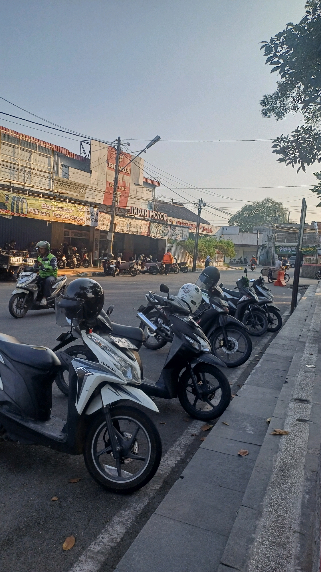 The image depicts a street scene in Klaten, Indonesia, focusing on a row of parked motorcycles along a sidewalk. In the foreground, several scooters are parked, with a white and black Honda scooter prominently displayed. A black helmet rests on a dark-colored scooter behind it. Further down the street, more motorcycles are visible, some with riders.
Behind the motorcycles, a building with signage for "INDAH MOTOR" is visible, suggesting a motorcycle dealership or repair shop. The building has a faded red and white color scheme. Various banners and advertisements are also displayed on the storefront. Overhead, power lines crisscross the sky, and a street lamp is mounted on a pole. The environment appears to be urban, with a paved road, sidewalk, and buildings lining the street. The sky is clear with a light haze. The overall impression is that of a typical busy street in a Southeast Asian city.