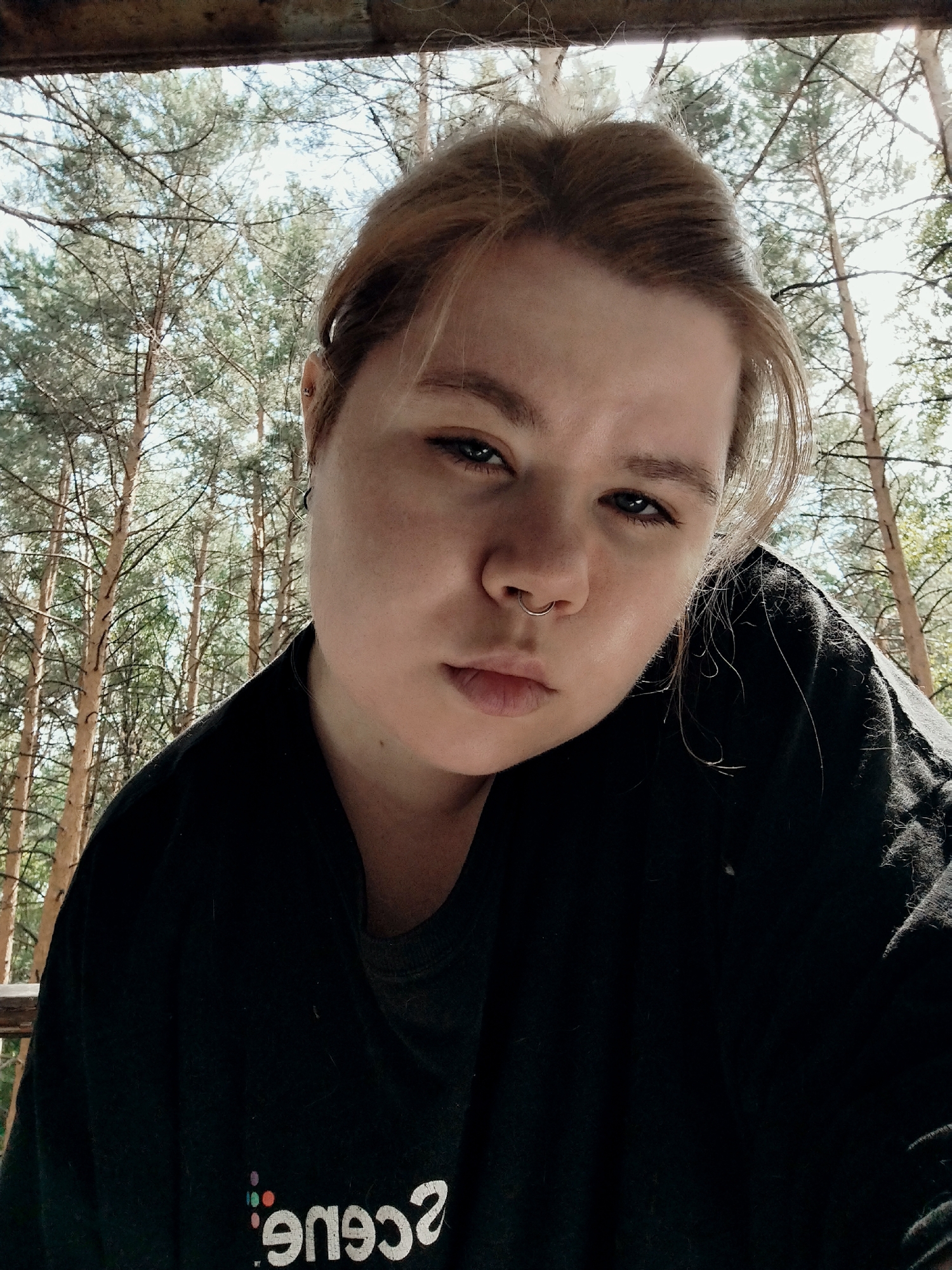 A close-up, low-angle selfie shows a young person with fair skin, light brown hair pulled back, and facial piercings including a septum ring and an earring. They are wearing a black t-shirt. The background consists of tall pine trees against a bright sky, suggesting an outdoor location, possibly a forest or wooded area. The lighting is natural, indicating daytime. The environment appears to be in a natural setting, with no discernible urban features.