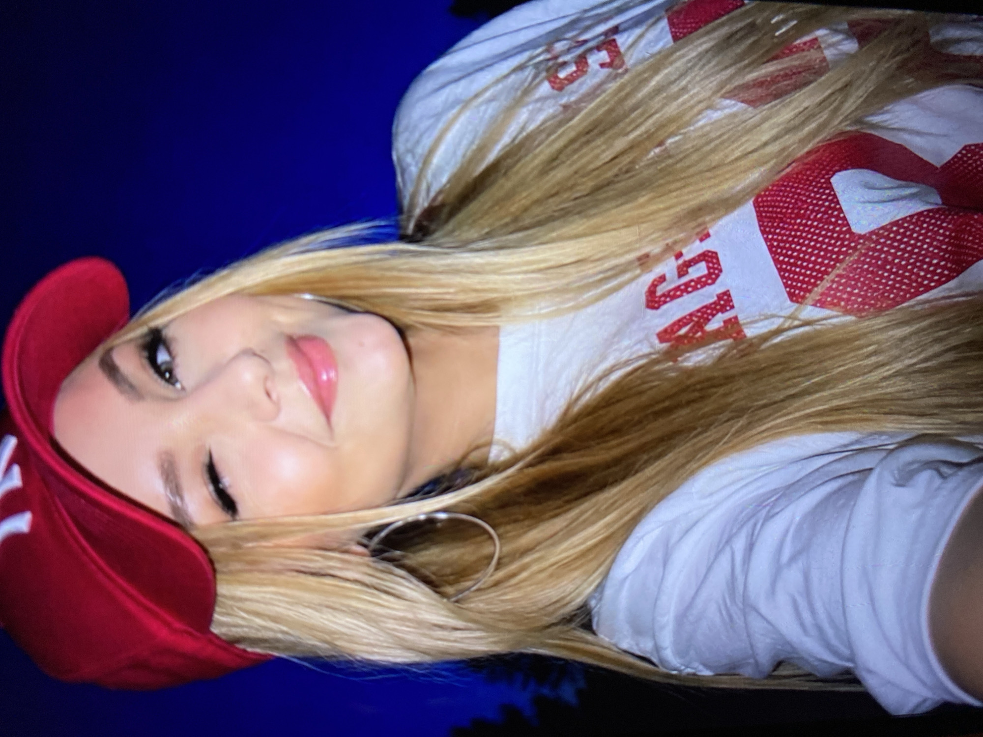 The image shows a close-up, slightly angled, selfie-style portrait of a young Caucasian woman with long blonde hair. She is wearing a red baseball cap with a white logo, a white t-shirt with red lettering and graphics, and large silver hoop earrings. Her right eye is closed in a wink, and she has pink lip gloss on. The background is a dark, blurry blue, with indistinct shapes suggesting trees or foliage. There is no indication that the image was taken in Luh, Ukraine, or any specific city.
