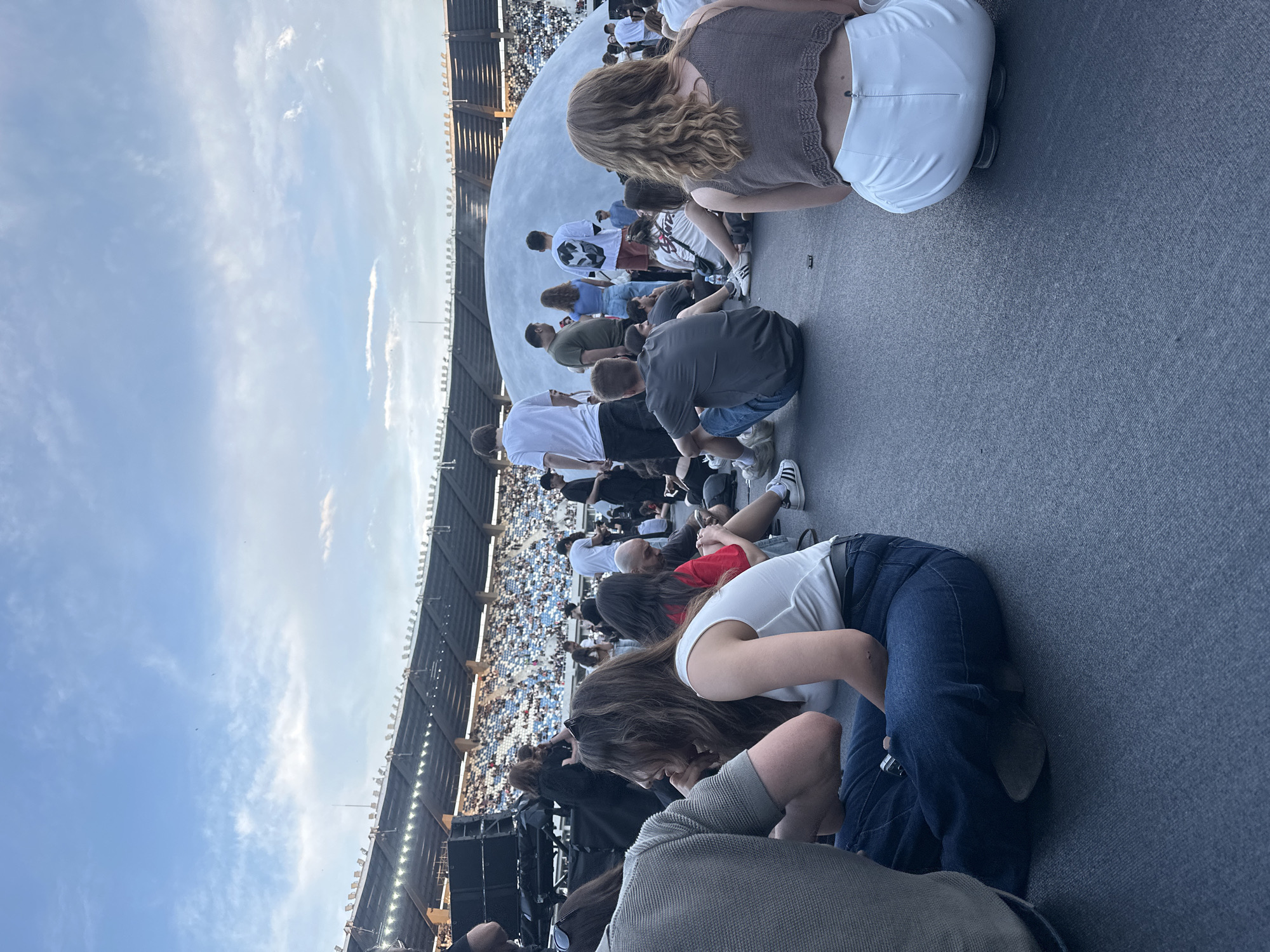 An outdoor concert is taking place at a stadium in Tbilisi, Georgia. The stadium seating is visible in the background, filled with spectators. In the foreground, a crowd of people are sitting on the ground, facing the stage area. A large, white, dome-shaped structure is prominent in the middle ground, and various stage equipment, including speakers, are also visible. The sky is partly cloudy, suggesting it is late afternoon or early evening. The people are dressed casually, in t-shirts, jeans, and shorts, indicating a relaxed atmosphere.